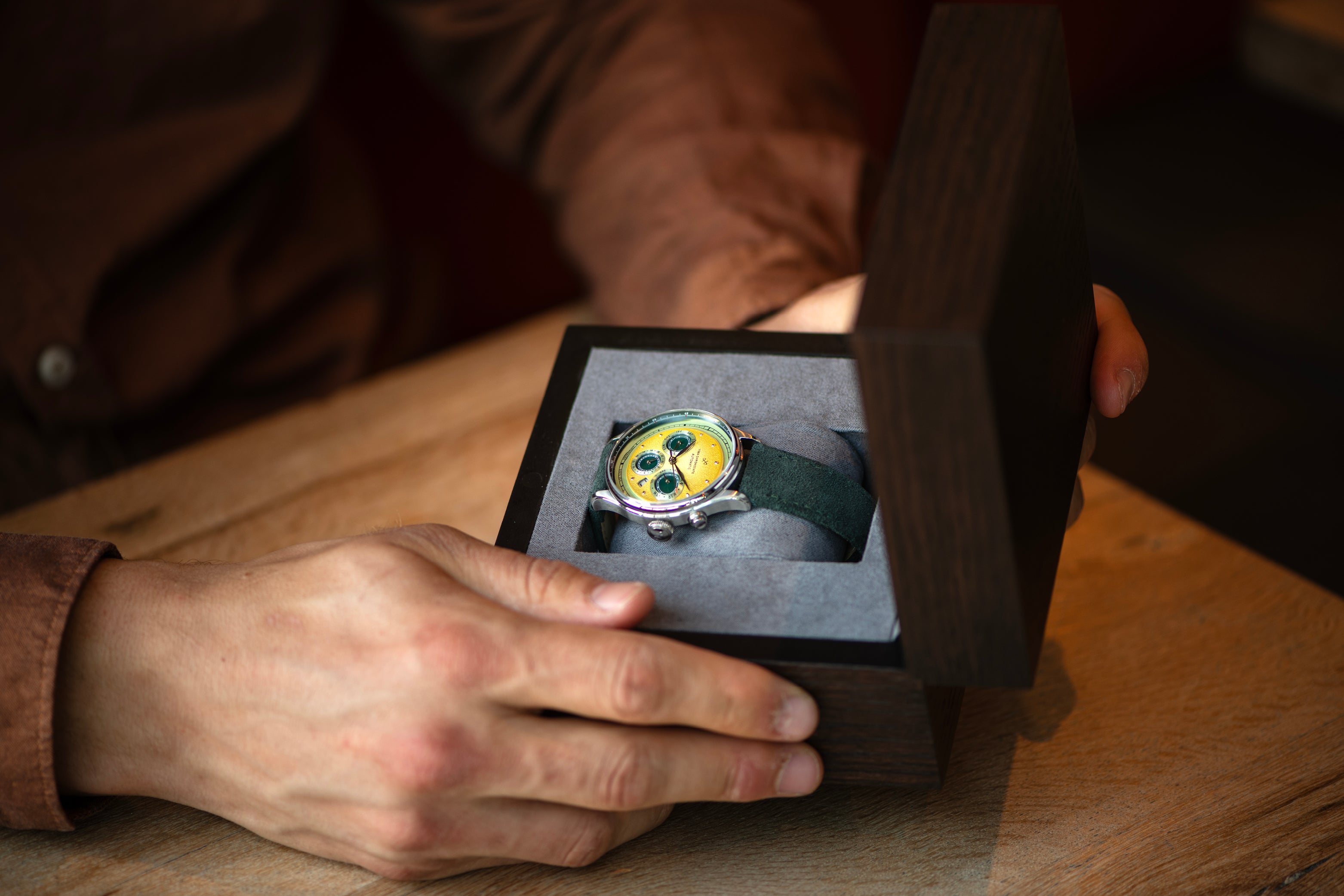 Man opening dark wooden box with wristwatch featuring yellow dial and dark fabric strap inside on wooden table