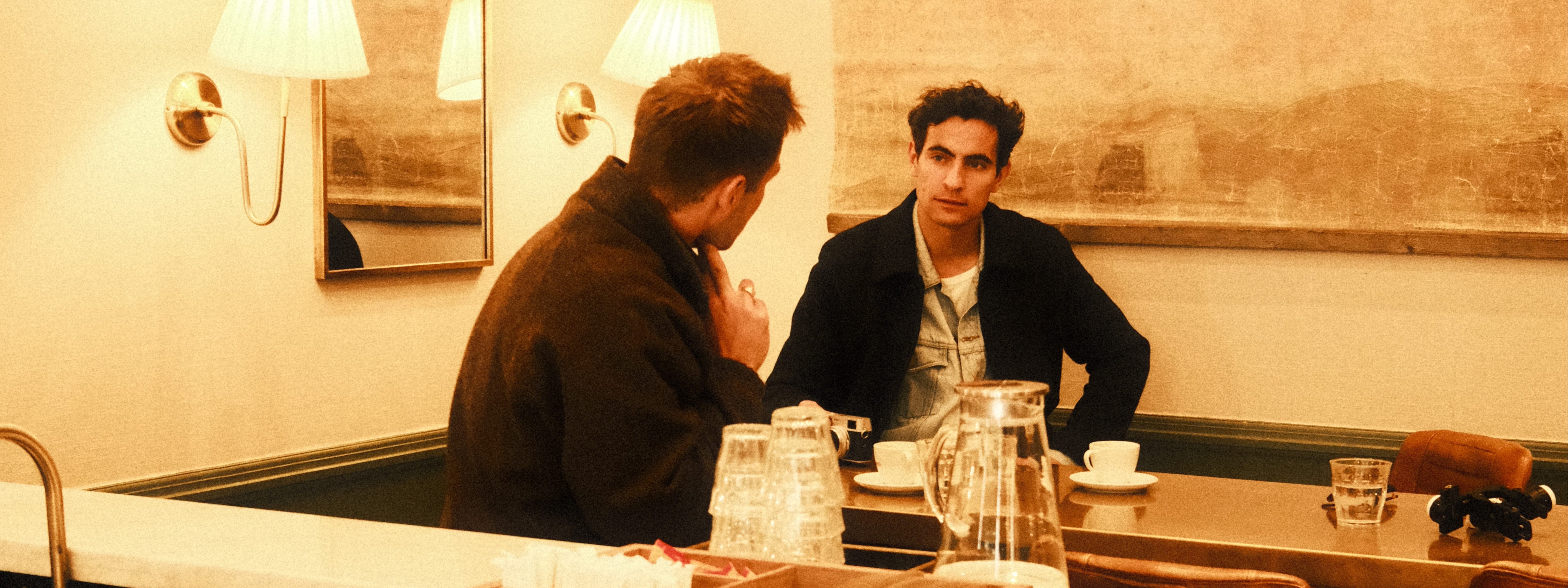 Two men engage in conversation seated at a corner booth in a warmly lit café, featuring soft beige walls, classic wall sconces, and a reflective mirror, creating an intimate and relaxed atmosphere with a subtle vintage European ambiance.