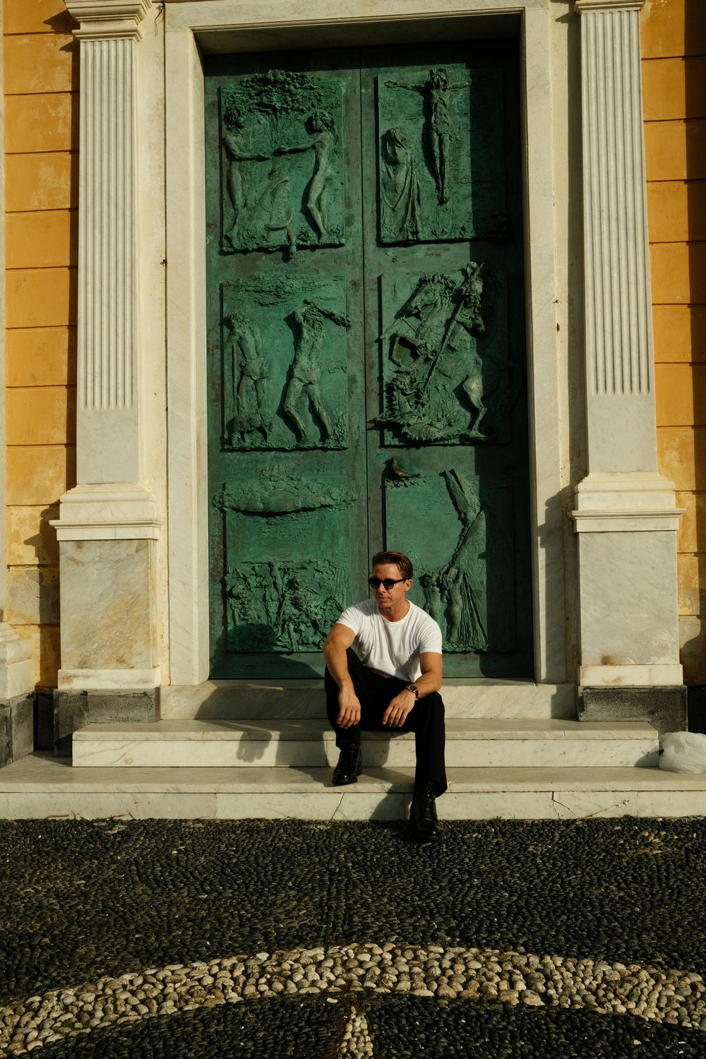 Man dressed in minimalist attire seated against ornate classical door, exuding relaxed Mediterranean style and timeless elegance wearing 1954 Amalfi Mechanical, Gold / Midnight Blue by AV86