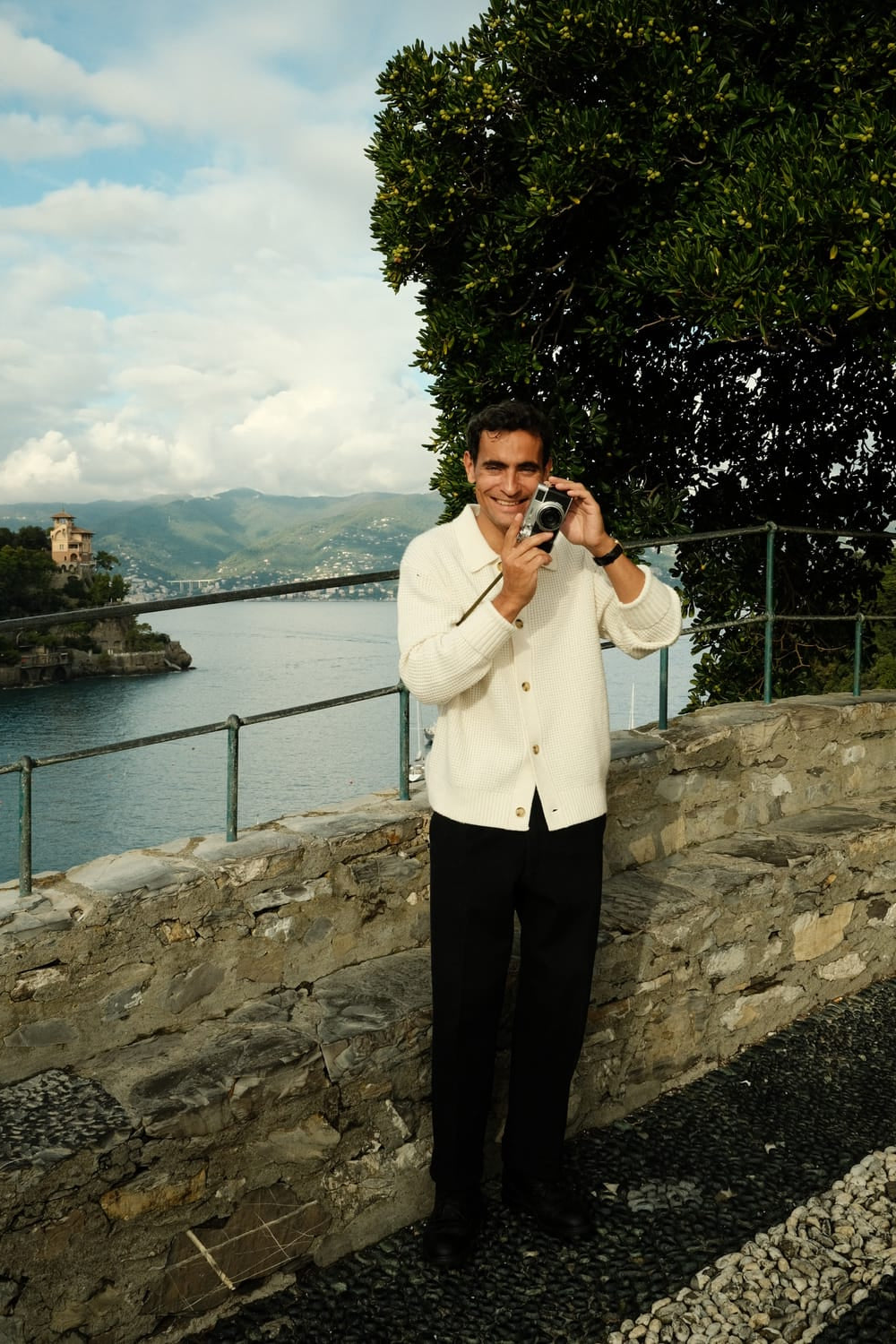 Man wearing 1954 Amalfi Mechanical, Steel / Off White by AV86, holding a camera near a scenic waterfront with stone railing.