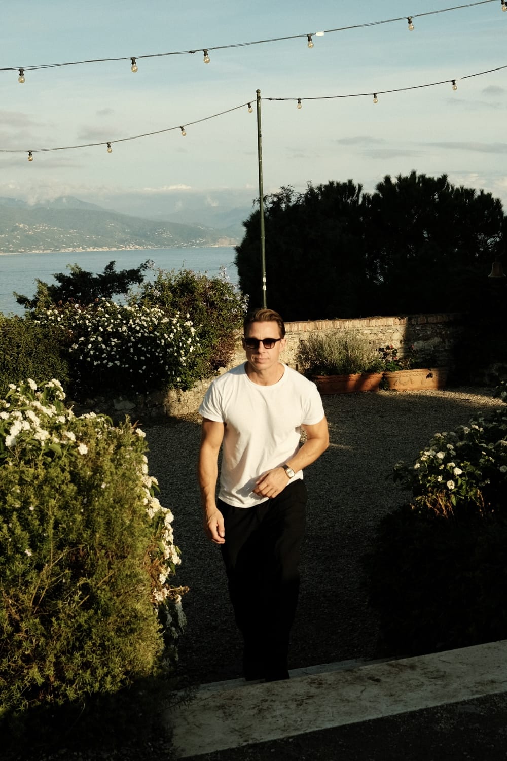 Man wearing sunglasses and a 1954 Amalfi Mechanical, Steel / Off White watch by AV86 walking outdoors near greenery and string lights.