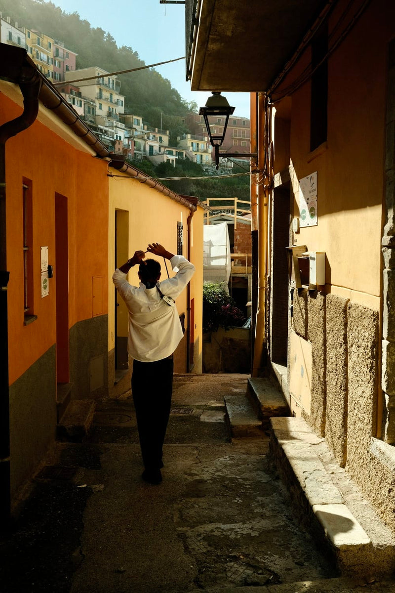 Person walking in a sunlit narrow street with colorful buildings, wearing 1954 Amalfi Mechanical, Steel / Off White by AV86.