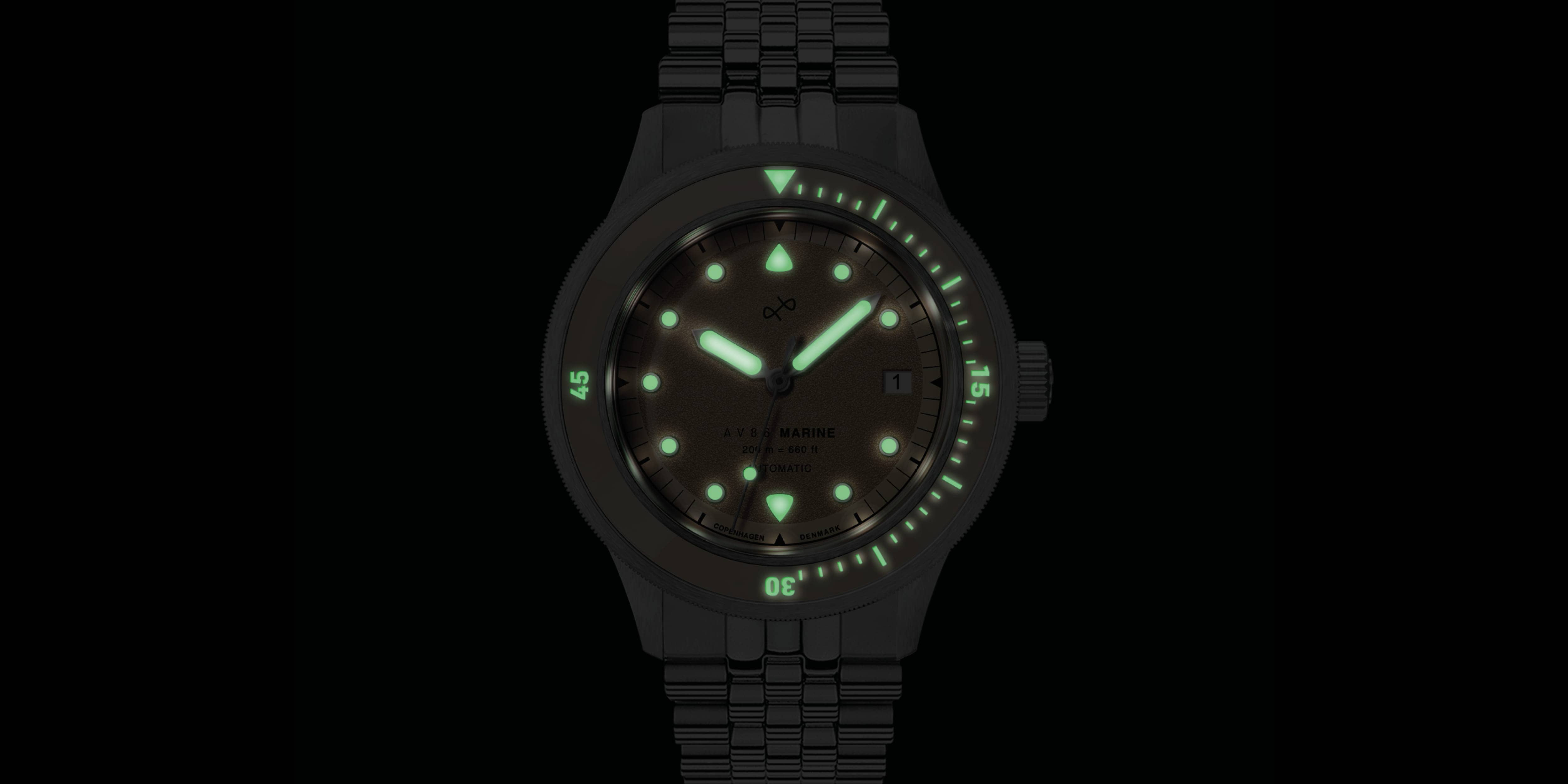 Dark background with a wristwatch showing luminescent green markers, hands, and bezel indicators on a metal bracelet.