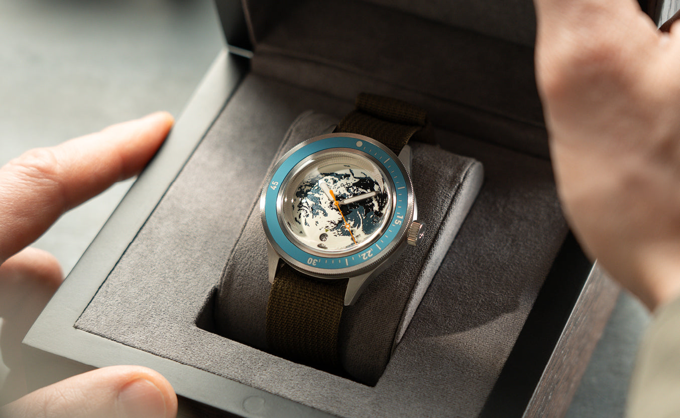 Luxury watch with turquoise bezel and globe-inspired dial, set in an open presentation box with a soft grey interior and olive fabric strap.