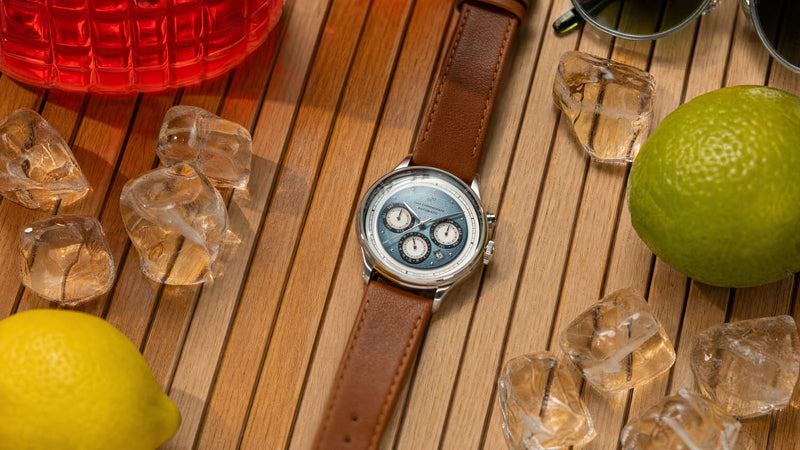 1564 Chronodate Automatic by AV86 features a vintage-inspired chronograph with sapphire crystal and leather strap, styled in a warm, casual setting with citrus fruits and ice cubes.
