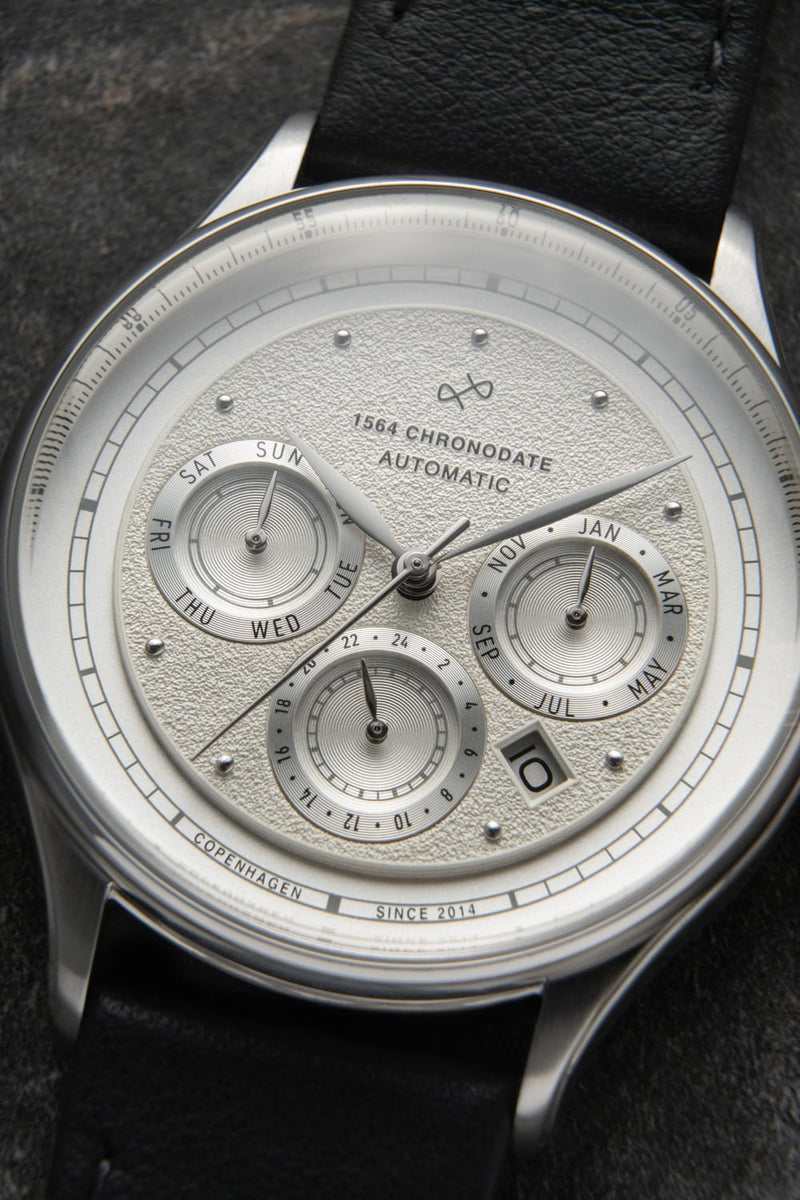 1564 Chronodate Automatic by AV86 exhibits a vintage-inspired automatic chronograph with a textured white dial, three subdials for day, month, and 24-hour time, sapphire crystal, polished stainless steel case, and a black leather strap, reflecting refined Danish design and Nordic minimalism.