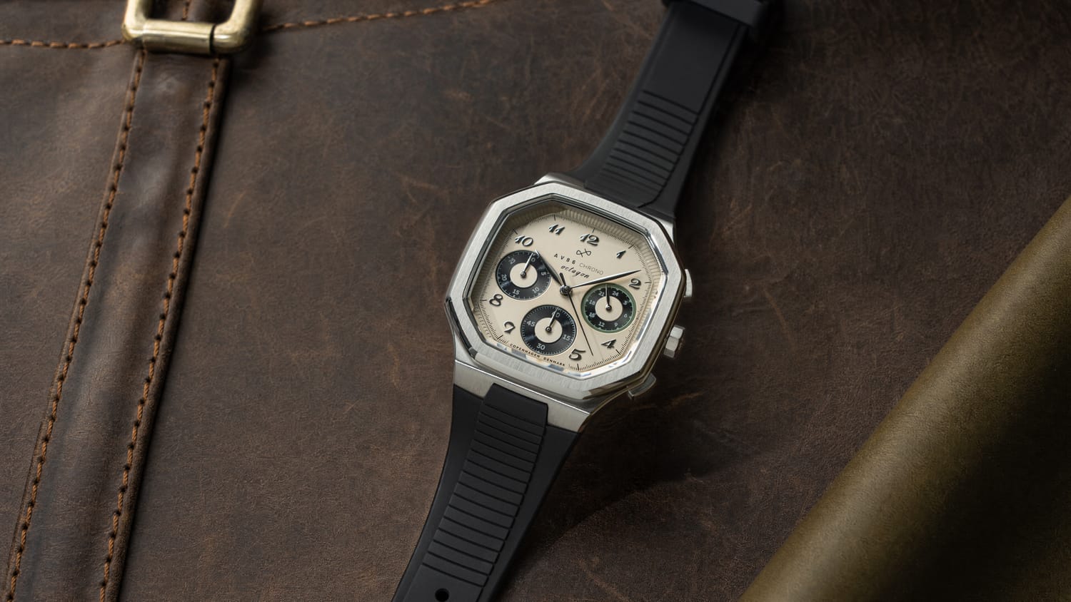 1904 Chrono, Steel / Off White & Black by AV86 featuring a stainless steel case, off-white dial with black chronograph subdials, black rubber strap, and sapphire crystal, resting on a dark brown leather surface evoking Scandinavian luxury and horological precision.