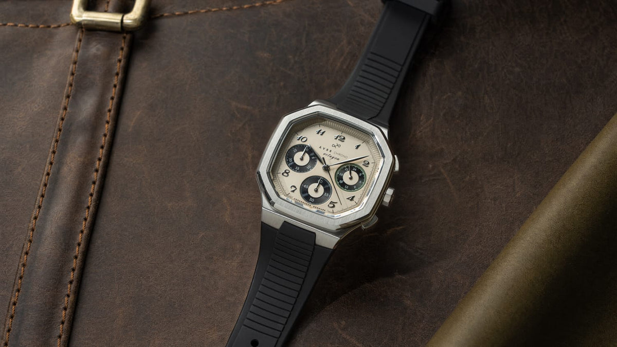 1904 Chrono, Steel / Off White & Black by AV86 featuring a stainless steel case, off-white dial with black chronograph subdials, black rubber strap, and sapphire crystal, resting on a dark brown leather surface evoking Scandinavian luxury and horological precision.