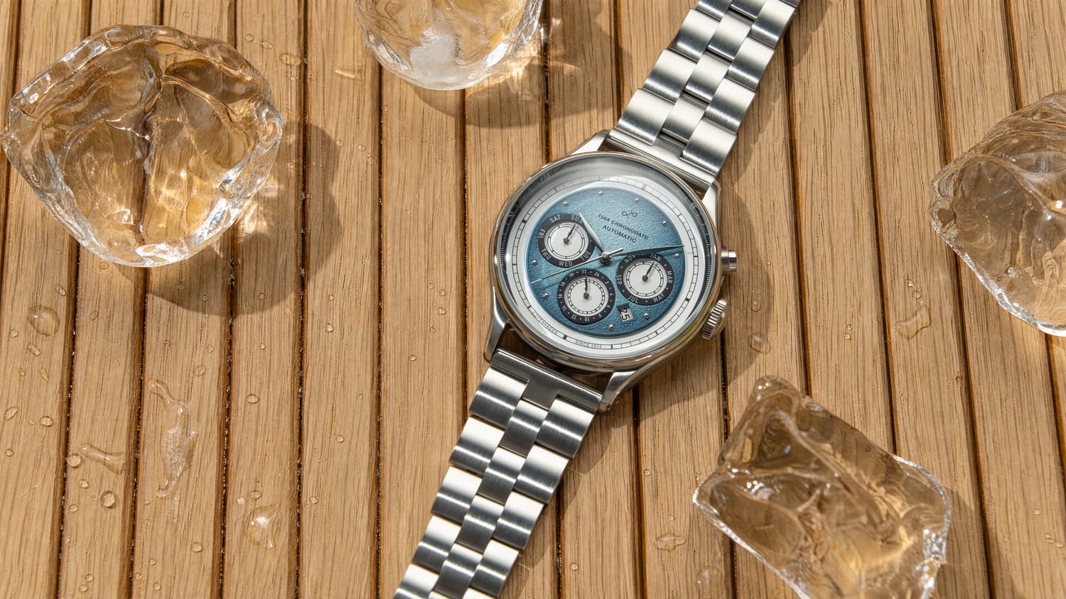 1564 Chronodate Automatic with a polished bracelet and ice blue dial featuring three chronograph subdials, presented by AV86 on a natural wood surface with melting ice cubes, conveying a blend of precision and Scandinavian minimalism.