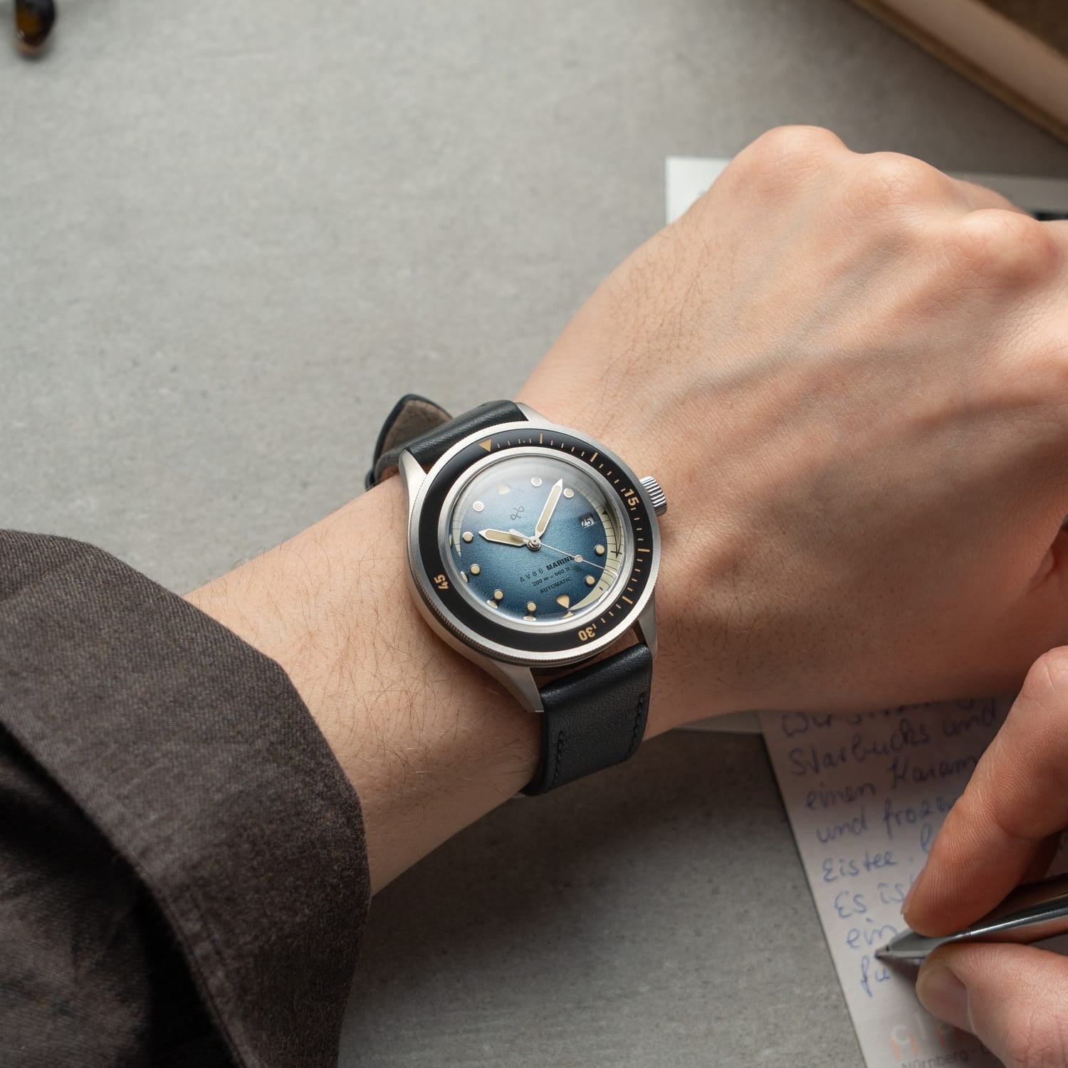 1956 Marine Automatic, Steel / Blue Fade wristwatch by AV86 with a blue fade dial and black leather strap on a wrist writing notes
