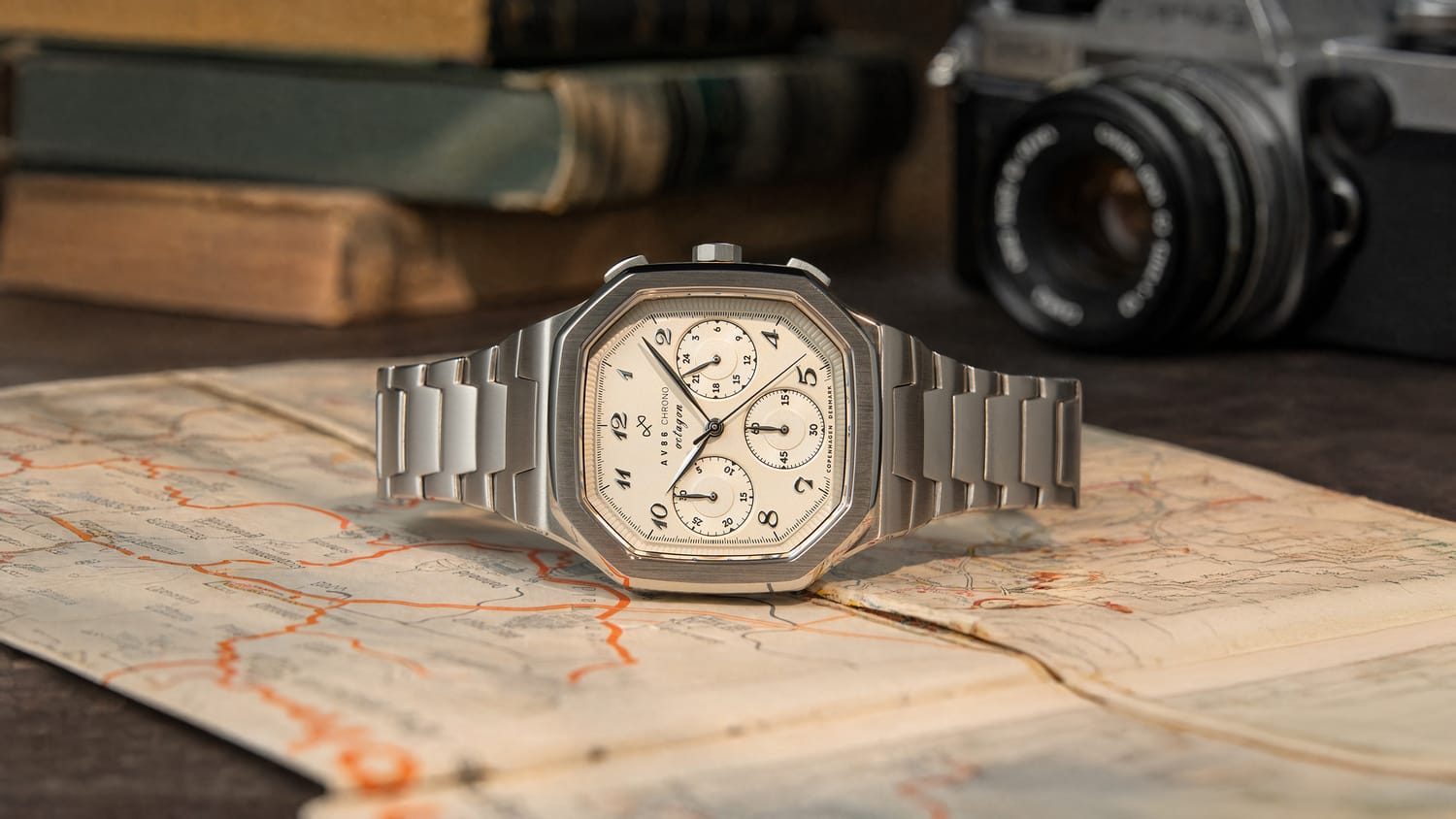 1904 Chrono by AV86, a luxury chronograph with a stainless steel bracelet, off-white dial, sapphire crystal, and vintage-inspired detailing, resting on a map with classic camera background.