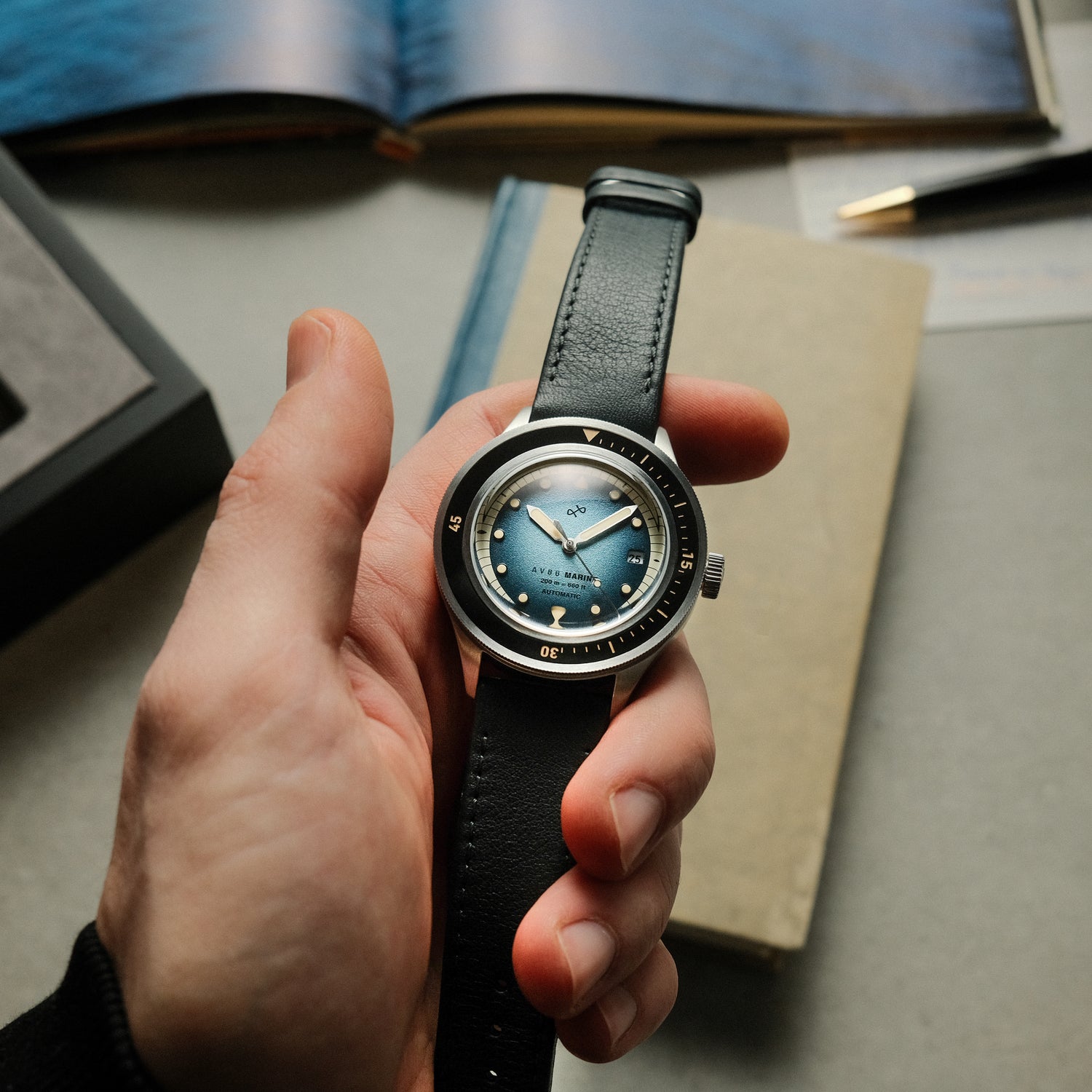 Hand holding 1956 Marine Automatic, Steel / Blue Fade with black leather strap against neutral background, by AV86 with vintage-inspired style