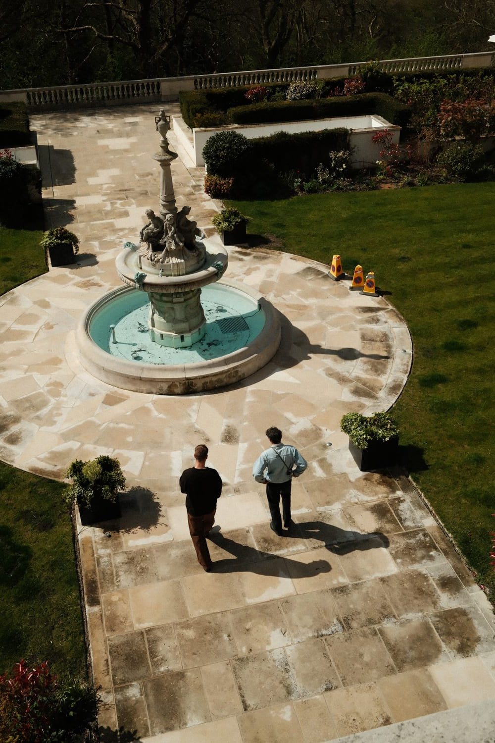 Two men walk along a sunlit stone pathway toward an ornate fountain in a manicured garden, evoking a serene and contemplative atmosphere that complements the refined elegance of the 1564 Chronodate Automatic by AV86.
