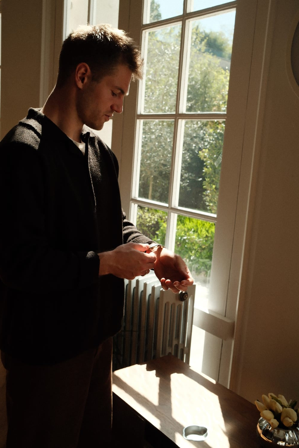 A man in casual dark attire adjusts the 1564 Chronodate Automatic by AV86 while standing near a sunlit window, surrounded by soft natural light and greenery, embodying a calm, minimalist Scandinavian atmosphere.