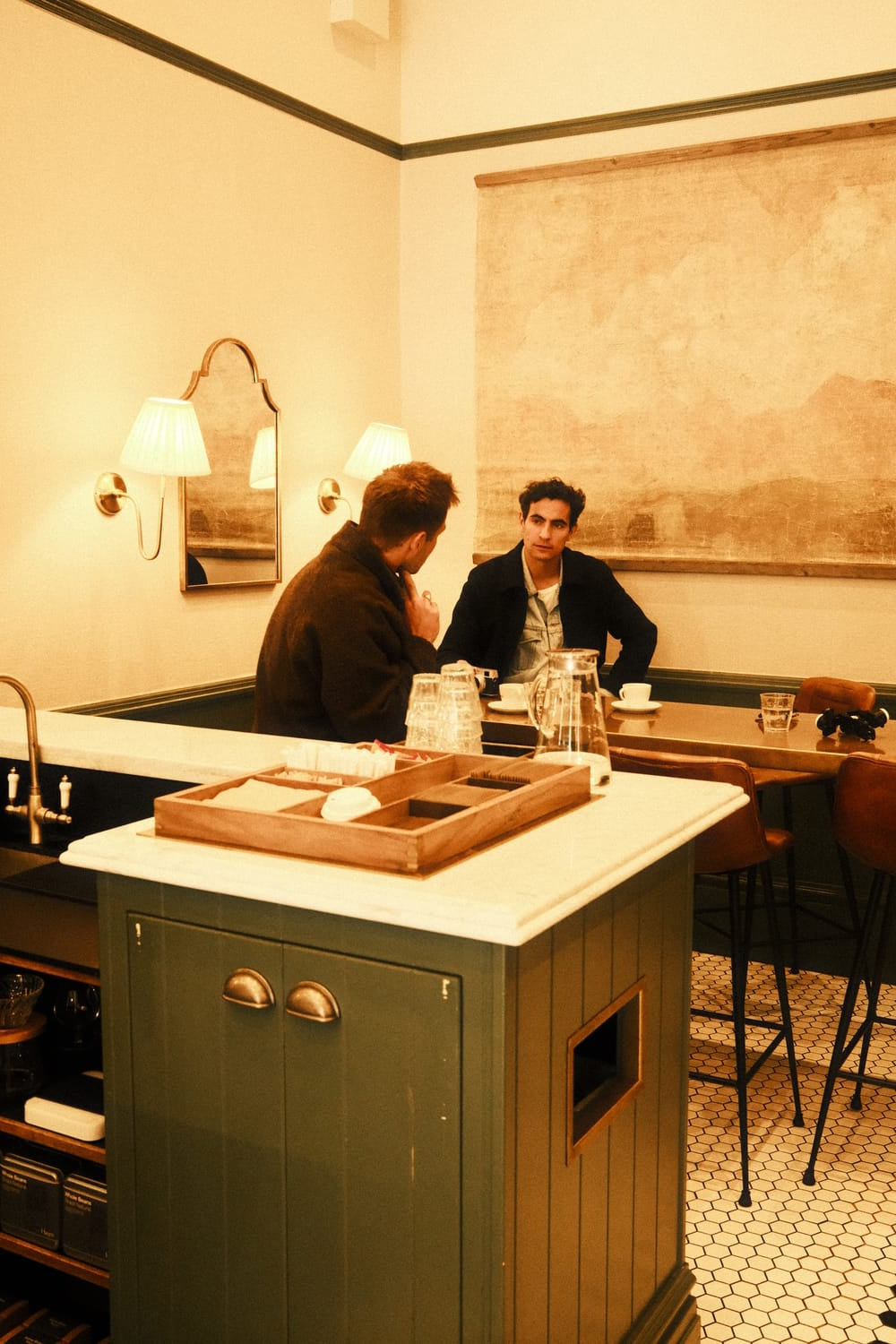 Two men engage in conversation at a minimalist café table, dressed in warm layers reflecting relaxed Scandinavian style, with subtle light accentuating a refined ambiance, evoking an intimate moment suited to the understated luxury of the 1564 Chronodate Automatic, Steel / Blue by AV86.