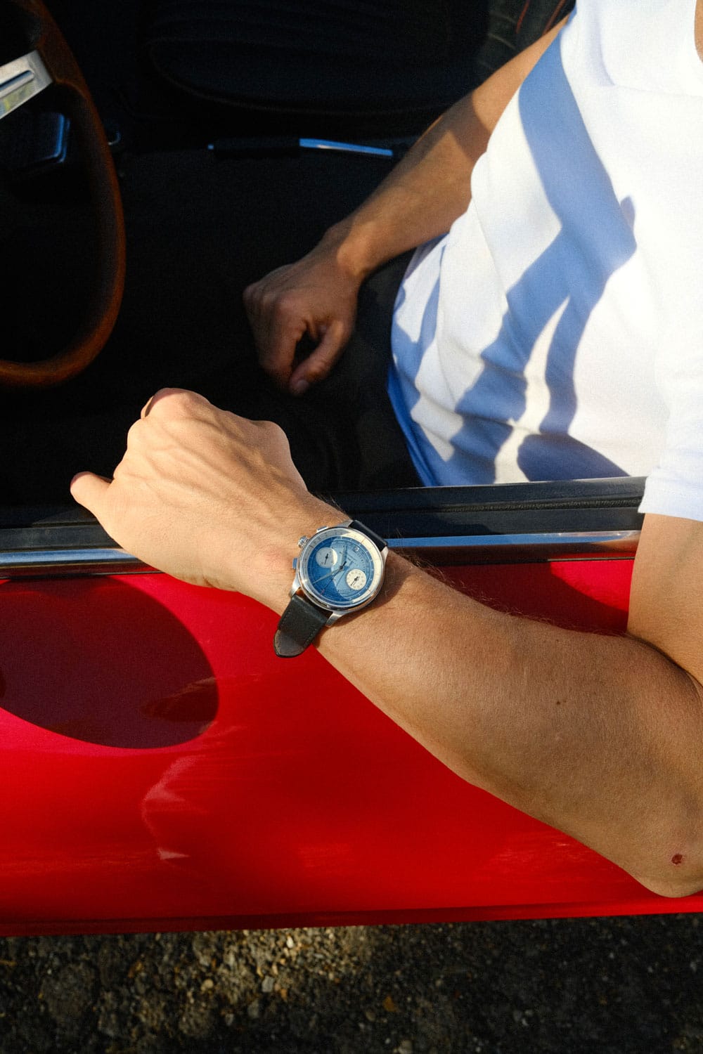 Wrist featuring an AV86 chronograph with a blue dial and subdials, polished case, and black leather strap, resting on the open window of a bright red vehicle, worn by a person in a white and blue striped shirt, highlighting relaxed luxury and Scandinavian design.
