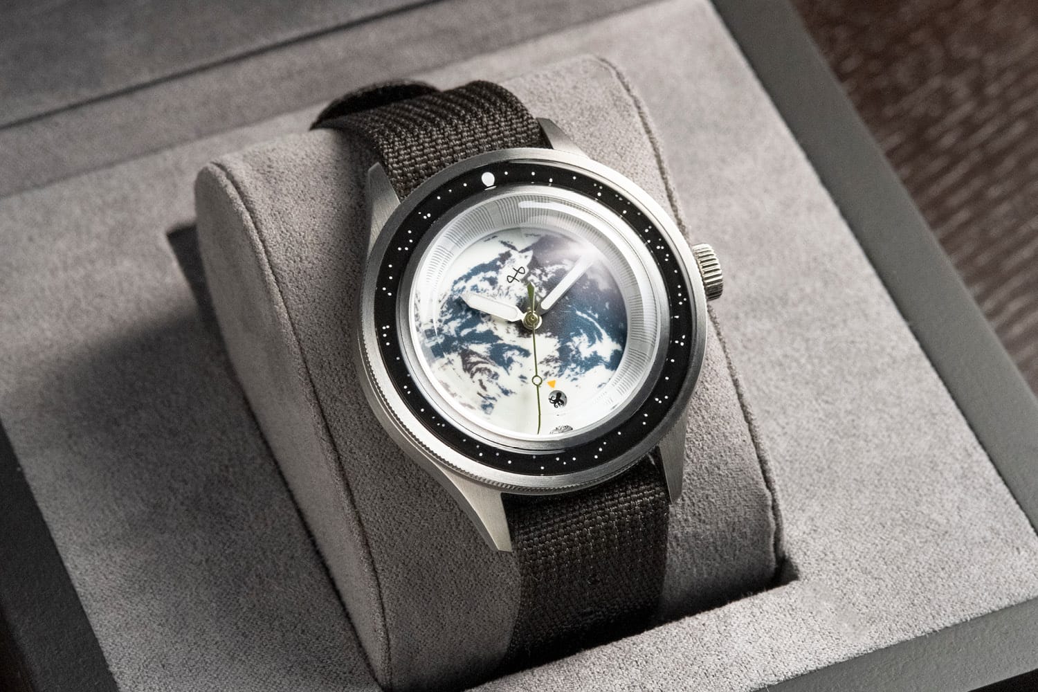 A close-up of a modern wristwatch resting on a soft gray display cushion inside a box. The watch has a dark woven fabric strap, a silver case, and a unique dial featuring an image of Earth viewed from space, framed by a black bezel with small white markers.