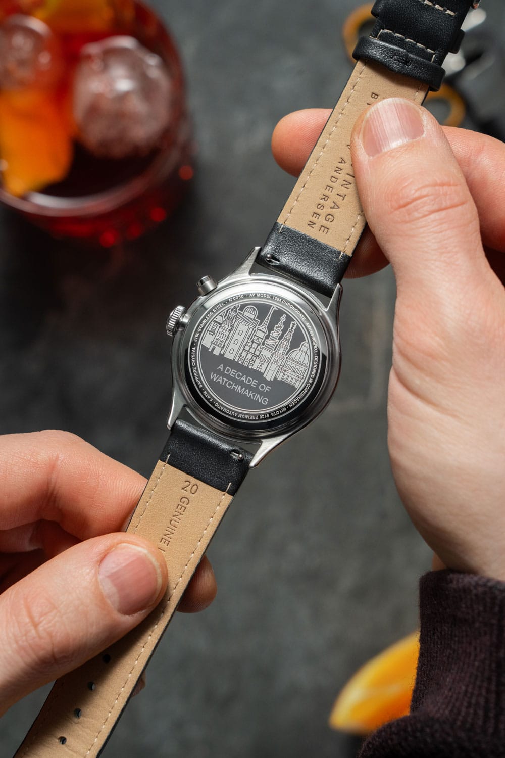 Close-up of a hand holding an AV86 watch showcasing engraved case back celebrating a decade of watchmaking with leather strap details