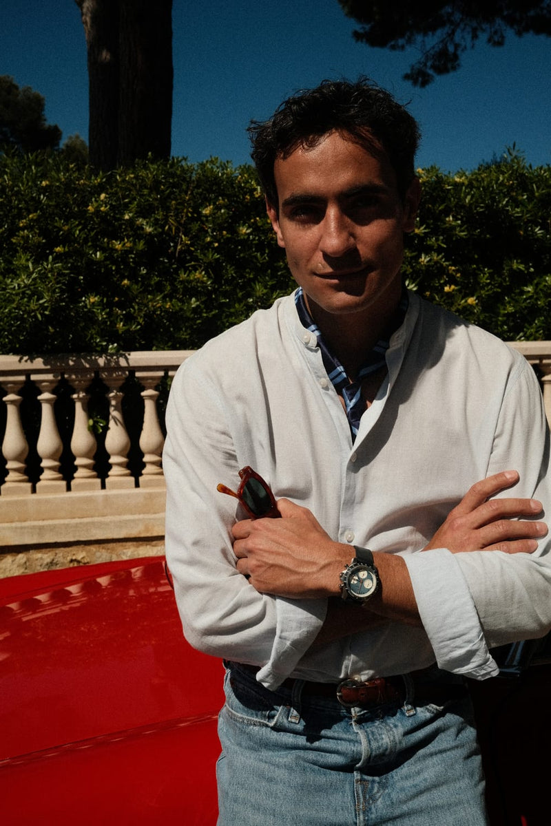 AV86 1960 Racing Chronograph, Steel / Sky Blue & Green quartz watch worn by a man in a white shirt beside a red classic car and greenery