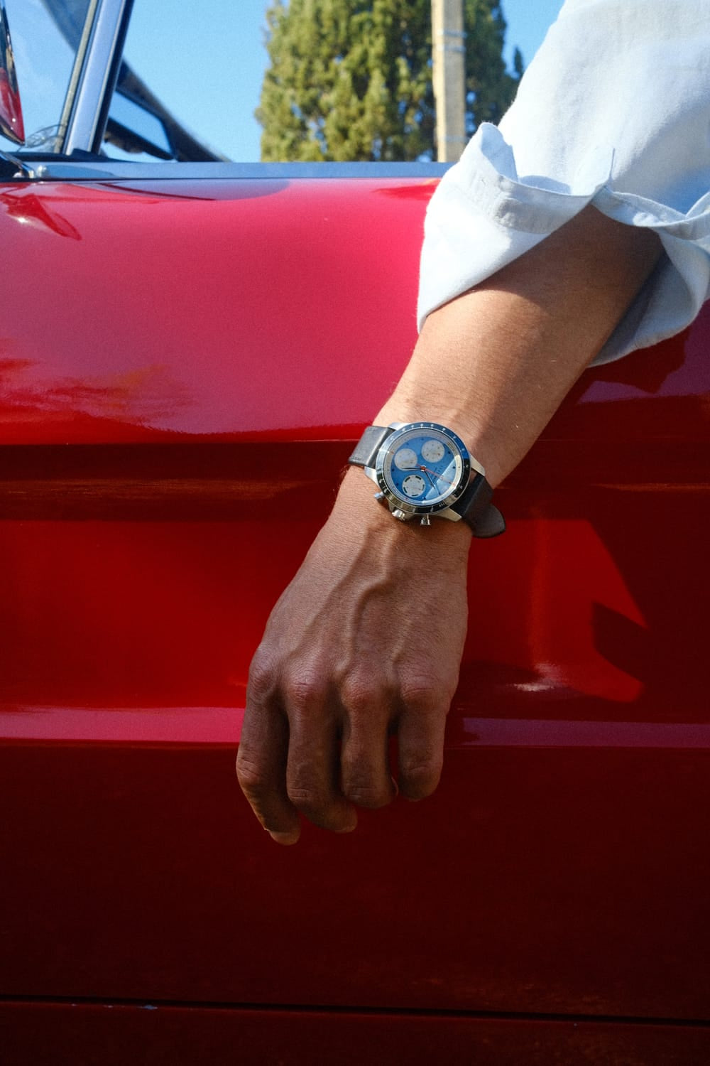 AV86 1960 Racing Chronograph, Steel / Sky Blue & Green watch with blue chronograph dial worn on a wrist beside a red classic car