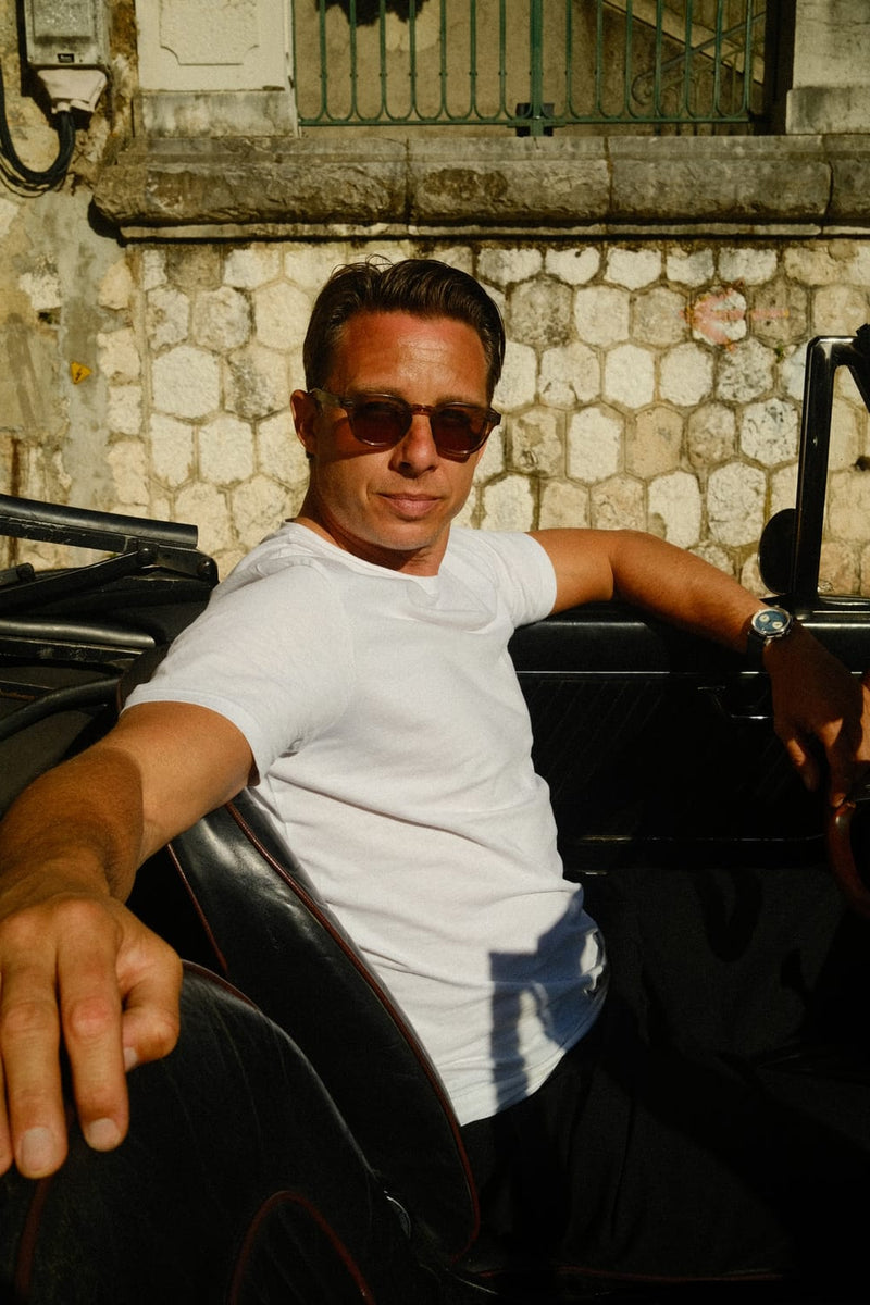 AV86 1934 Multichron, Steel / Sky Blue chronograph worn by a man in sunglasses and a white shirt beside a vintage car in warm sunlight