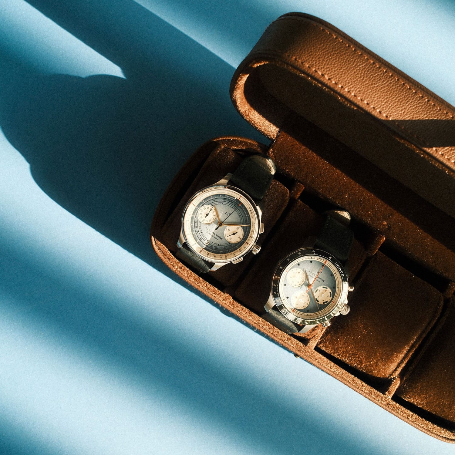Two vintage-inspired AV86 chronograph watches with dual subdials and sapphire crystals, featuring textured leather straps displayed in an open brown case on a soft blue backdrop, reflecting refined Danish design and minimalist horological craftsmanship.