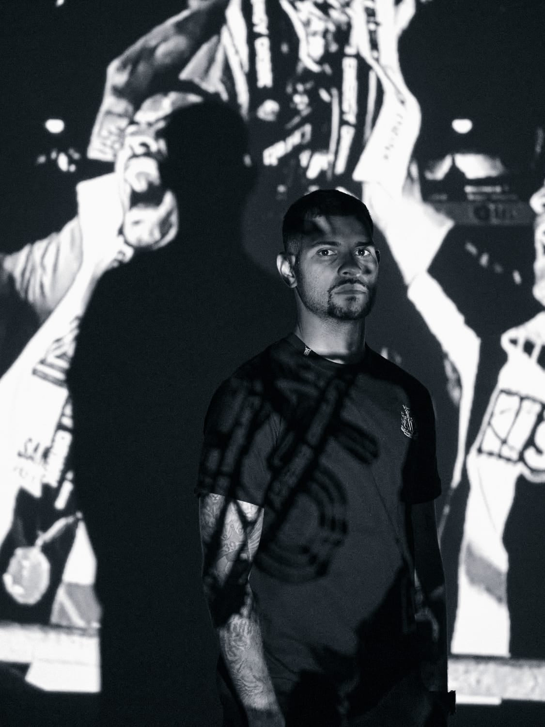 A man stands in a dimly lit setting with a dynamic black and white projection of football celebration behind him, showcasing a mood of triumph and focus linked to the Official Newcastle United Memorabilia Watch, Carabao Cup winners 24 / 25 by AV86.