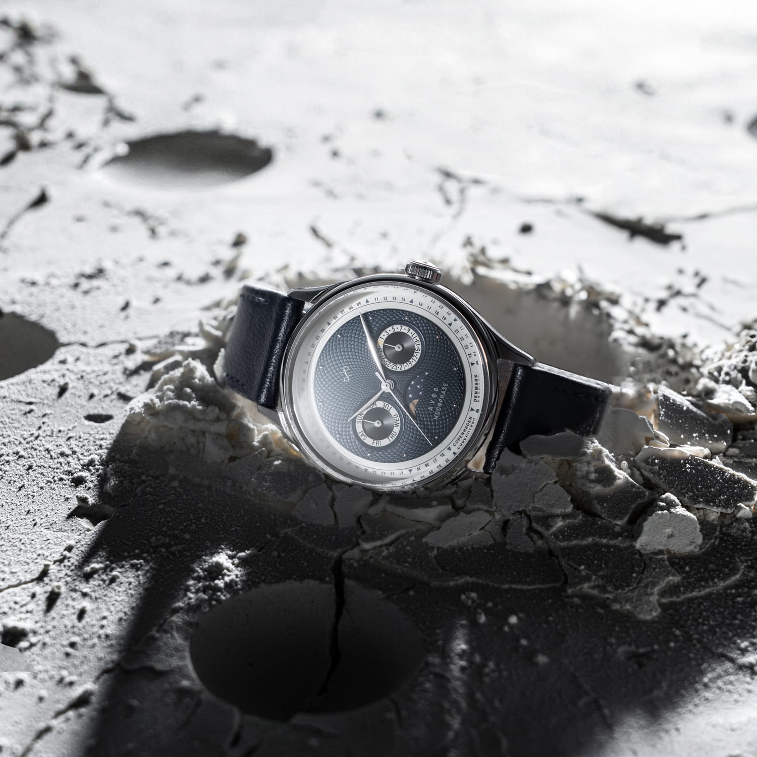Luxury wristwatch with black dial and leather strap resting on a textured surface resembling the moon's cratered landscape.