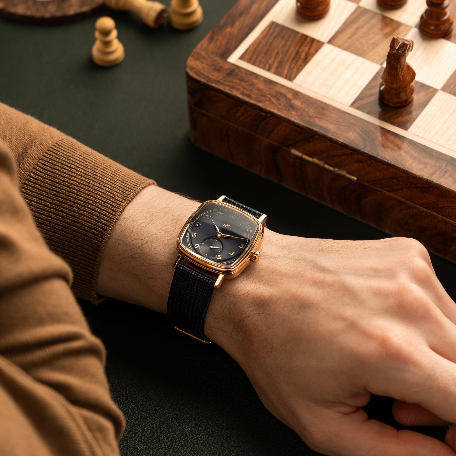 Wrist featuring a gold-toned square watch with black dial, vintage Arabic numerals, and textured black leather strap near a wooden chessboard.