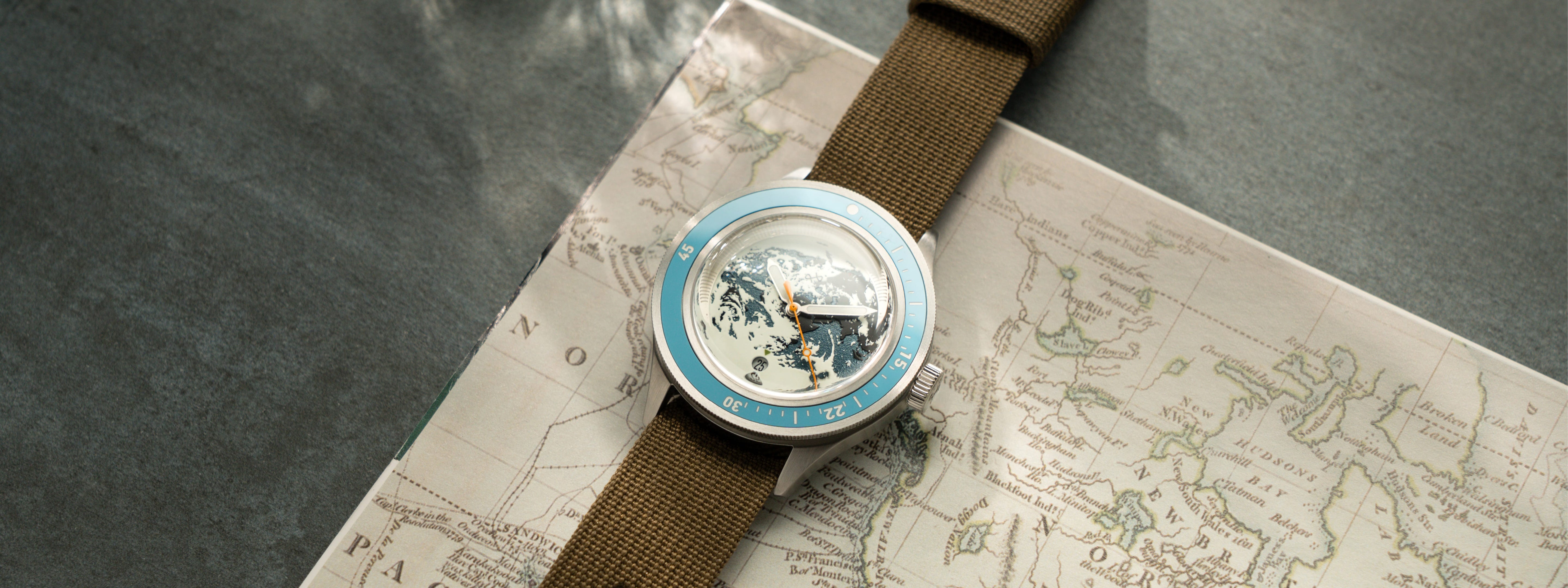 Wristwatch with light blue bezel, detailed globe dial, and olive green fabric strap resting on a vintage map background.
