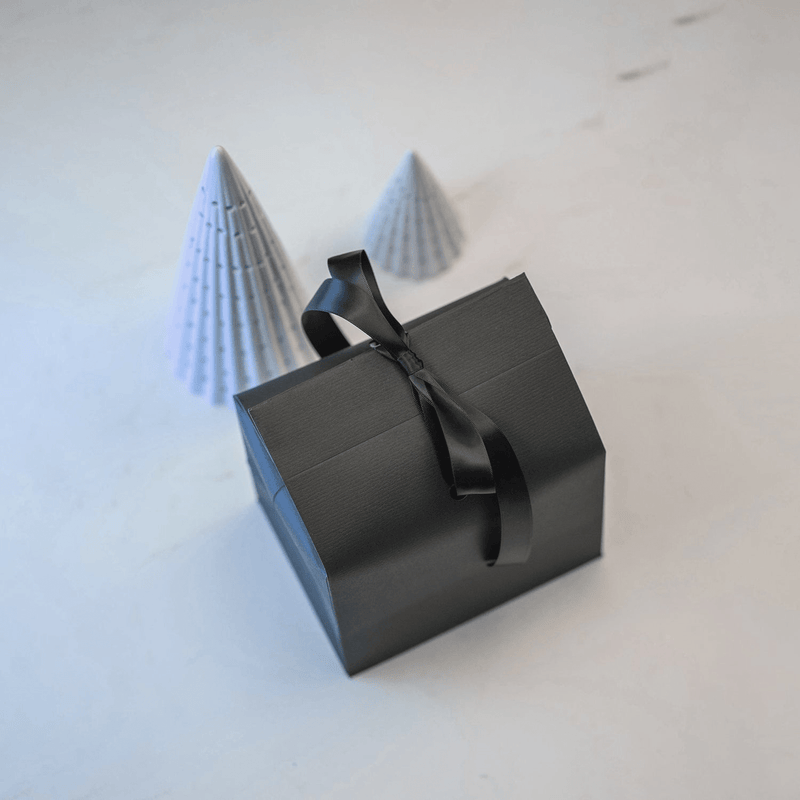 Emballage cadeau signé AV86, composé d'un élégant coffret noir orné d'un ruban de satin, accompagné de deux cônes en papier au style minimaliste posés sur un fond blanc.