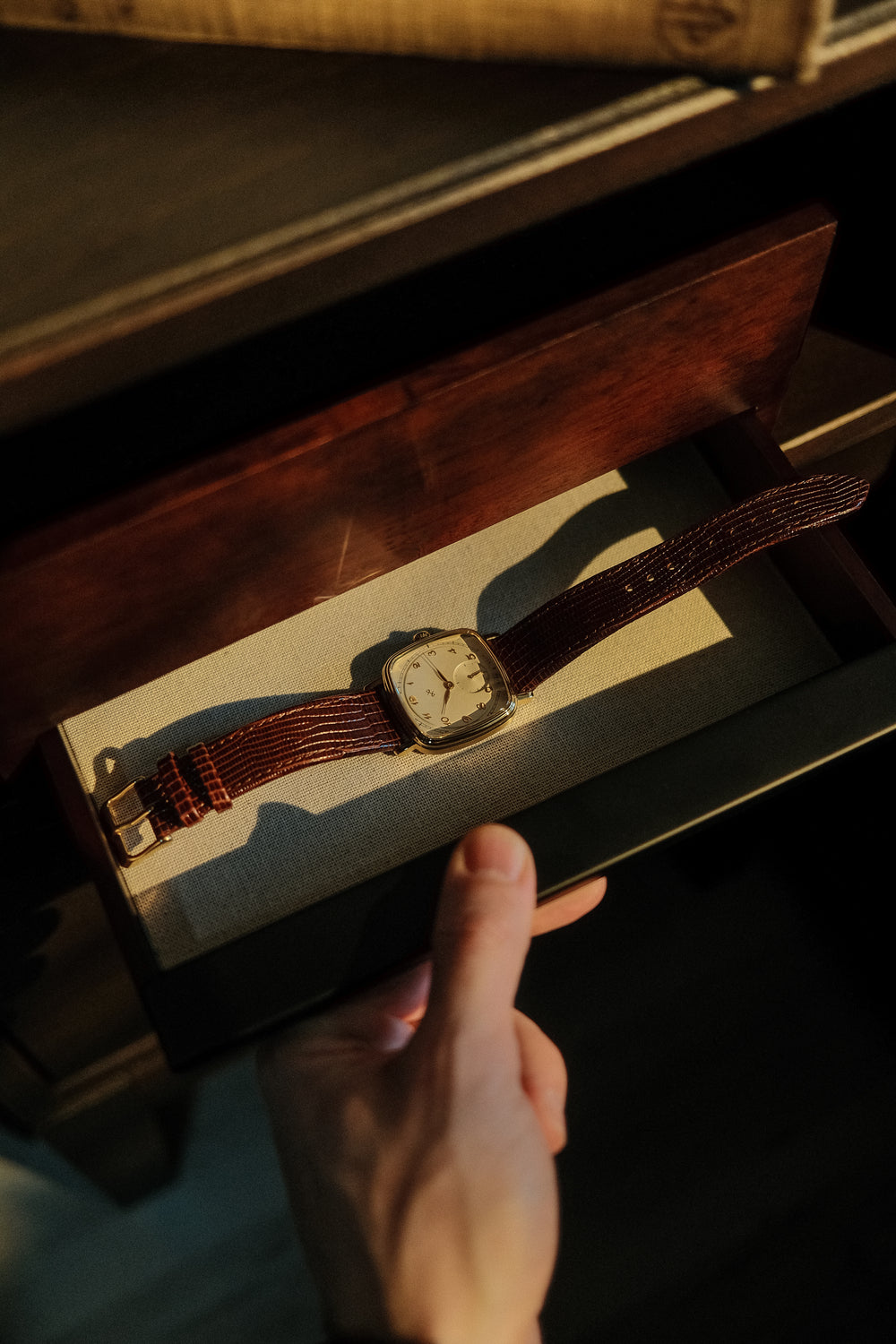 Hand holding open a wooden box with a vintage-inspired AV86 dress watch featuring cushion case and textured leather strap inside sunlight.