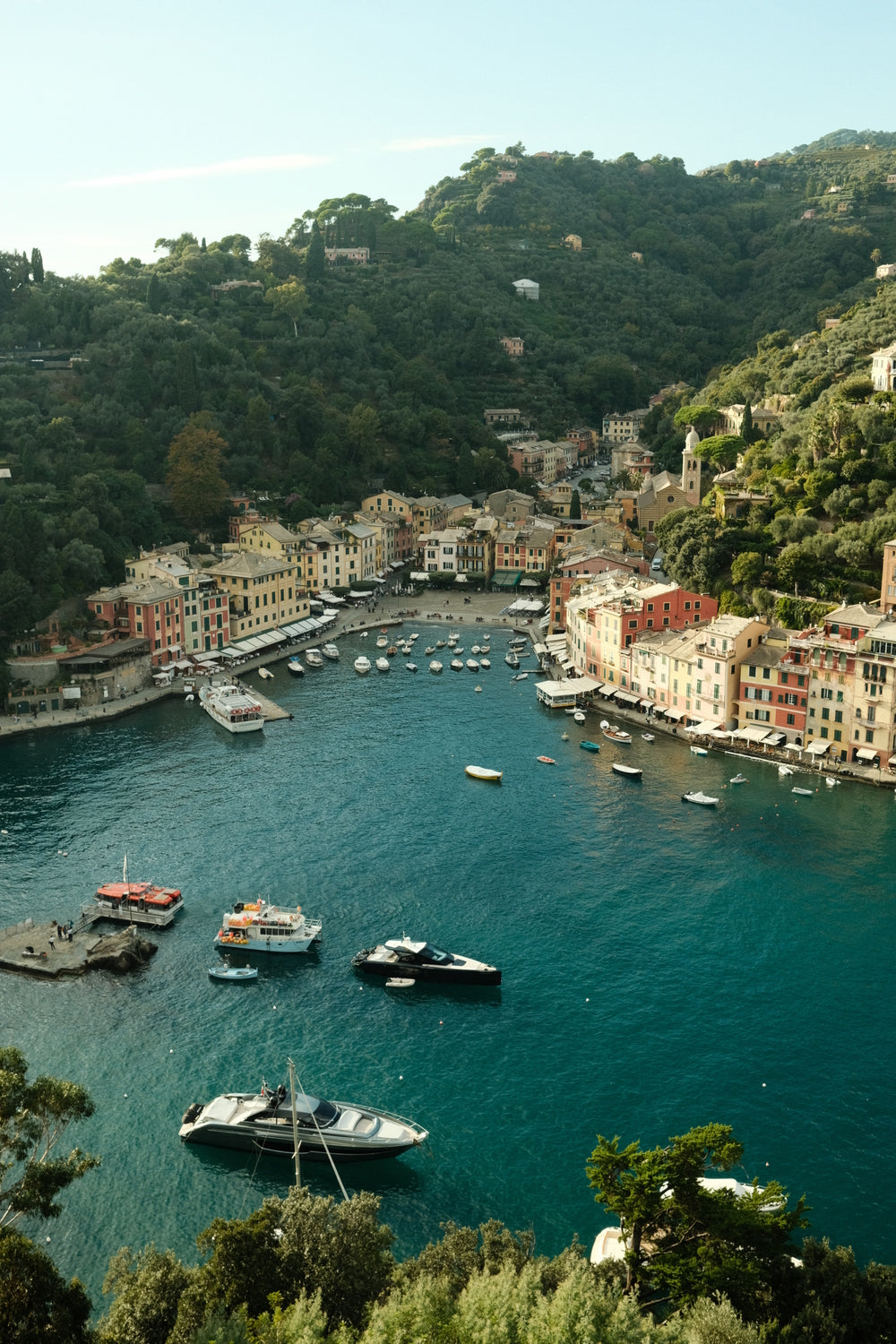 Coastal village with colorful buildings, lush green hills, and boats anchored in a tranquil Mediterranean harbor.