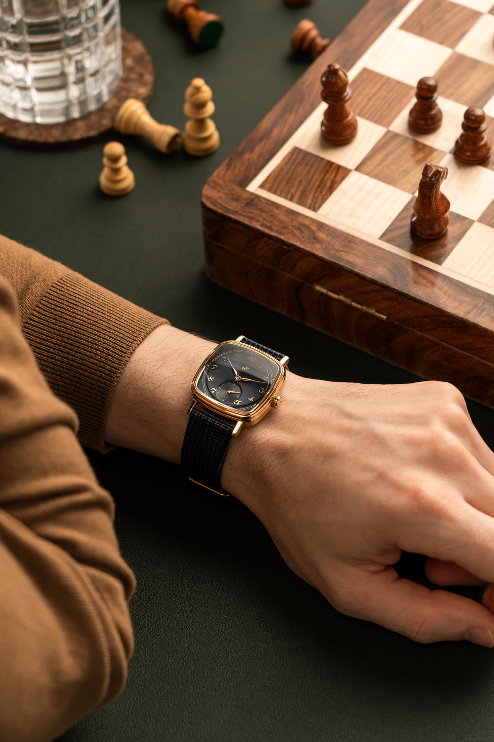 Wrist wearing AV86 dress watch with rounded square case, mechanical small seconds, sapphire crystal, and textured leather strap near chessboard.