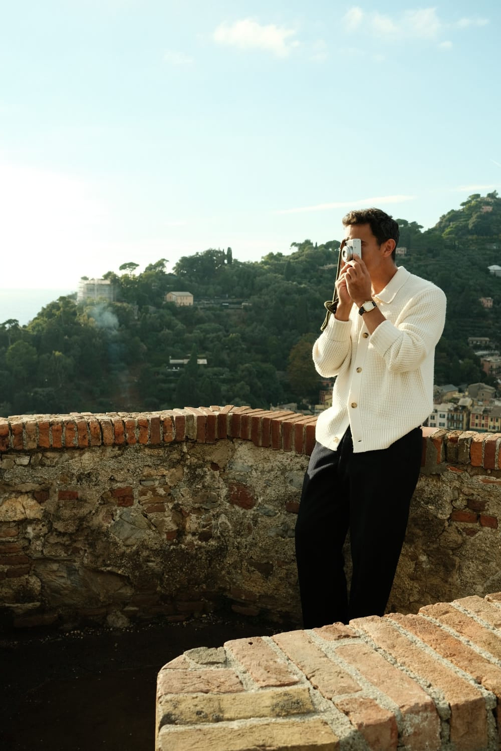 Man in cream knit cardigan and dark pants photographing coastal hillside village from rustic stone terrace in soft sunlight