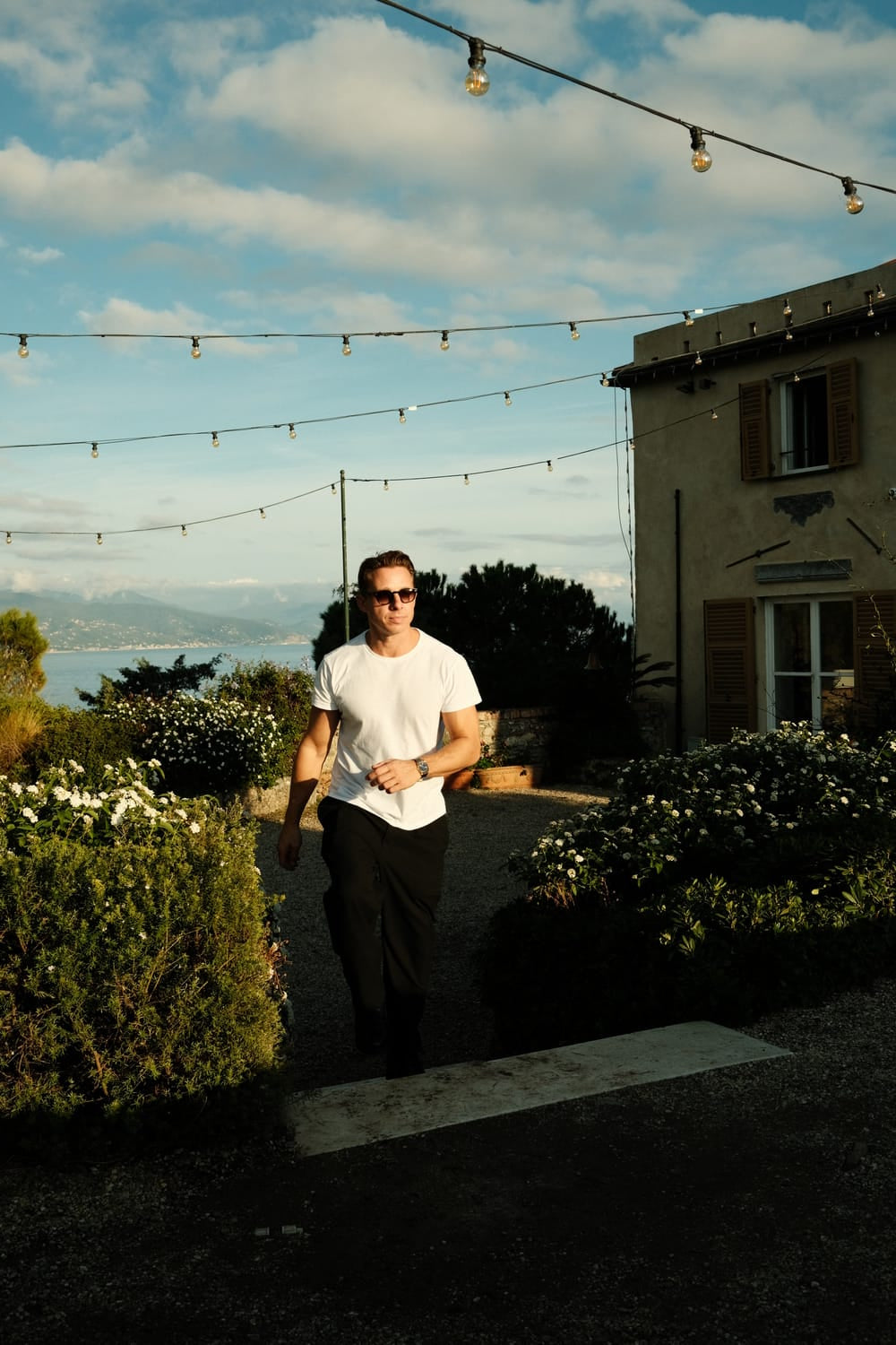 Man wearing sunglasses and wristwatch walking through a garden path near a rustic house under string lights at sunset