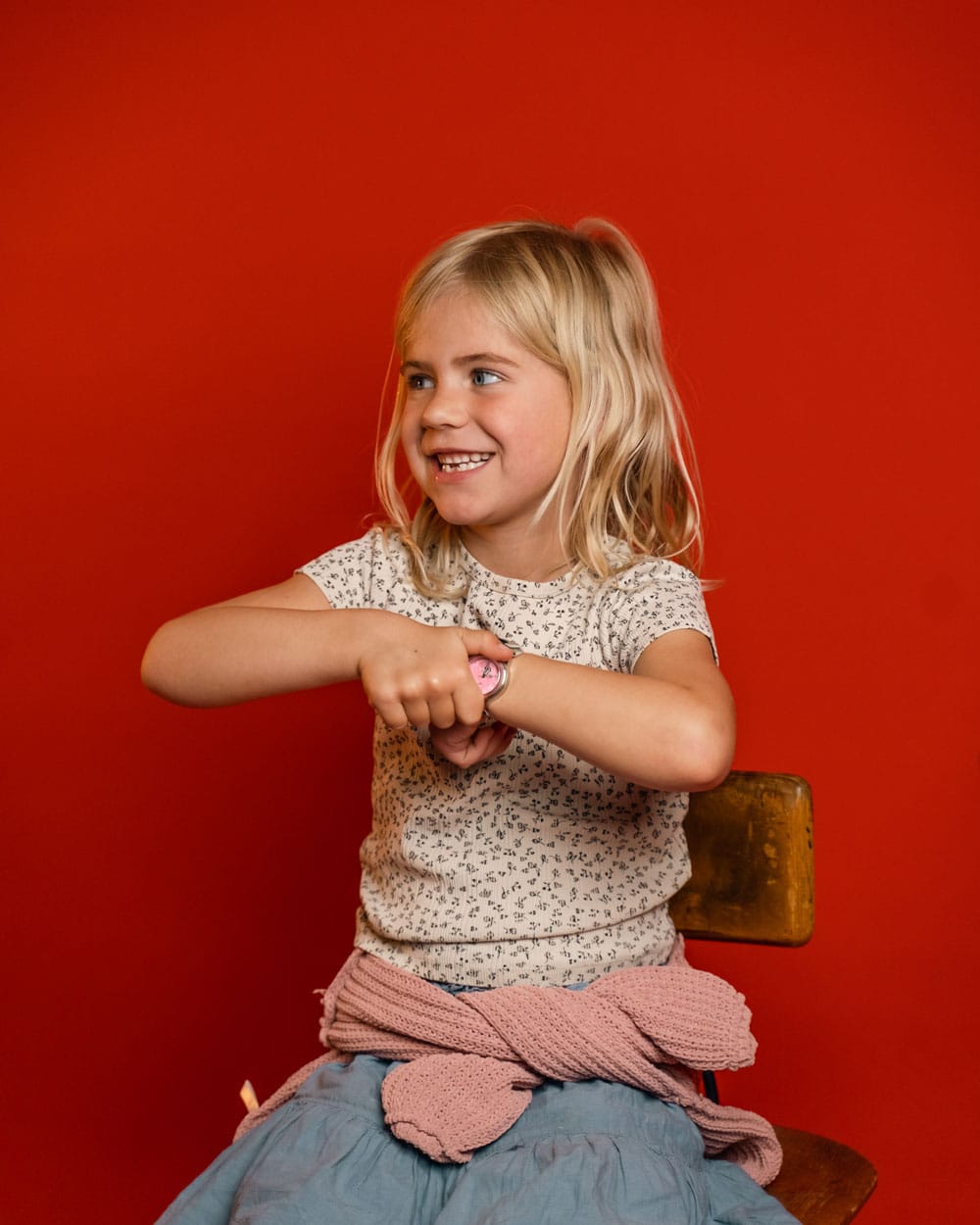 Young girl smiling in a patterned shirt and pink sweater, seated against a vivid red backdrop while adjusting a wristwatch