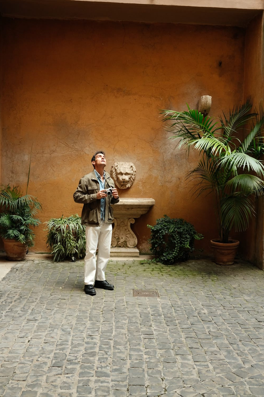 Man in casual jacket and white pants holding camera, standing on cobblestone courtyard with potted plants and stone lion sculpture.