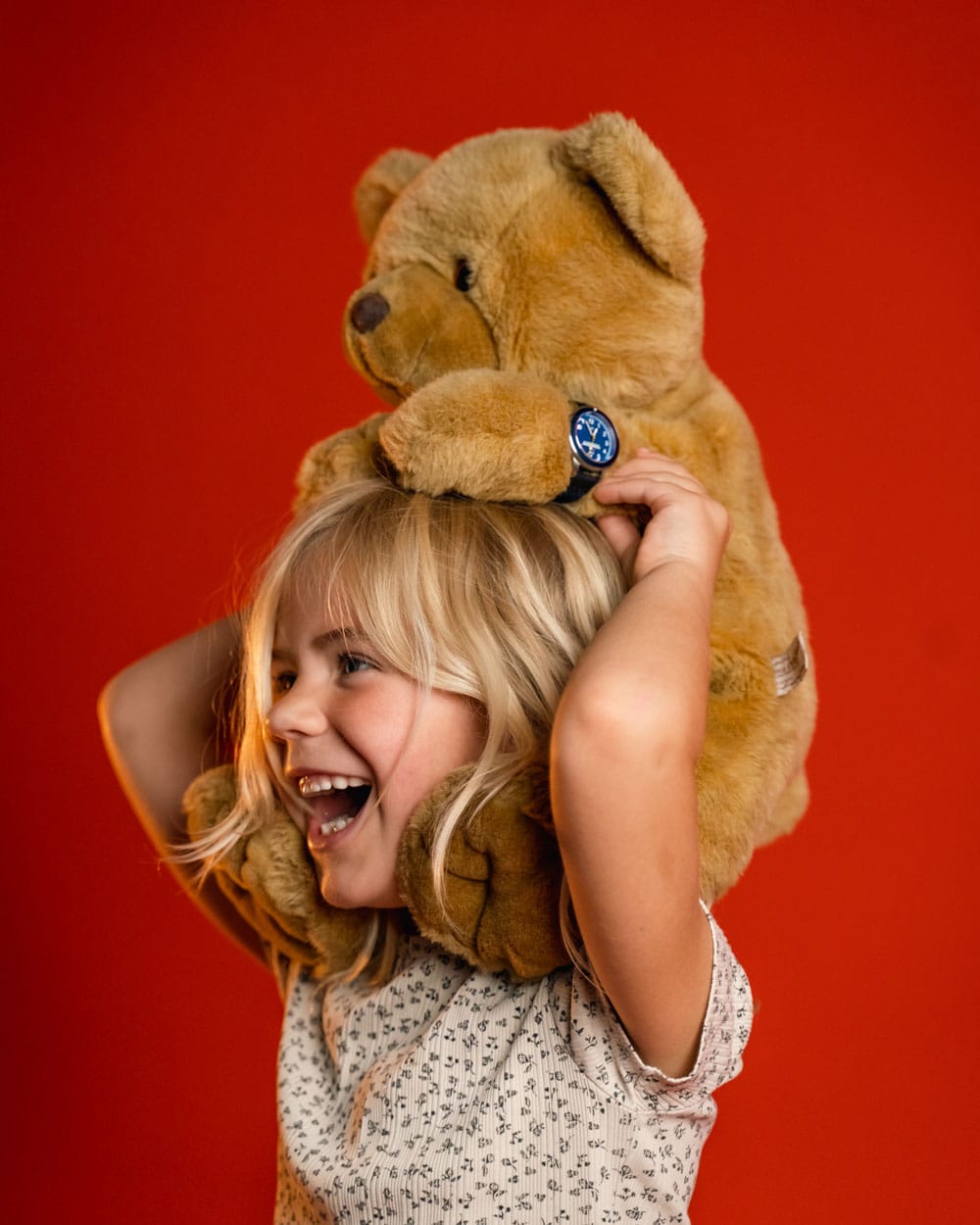 Smiling young girl in a patterned shirt carrying a large teddy bear on her shoulders, with a blue-dial wristwatch against a red backdrop