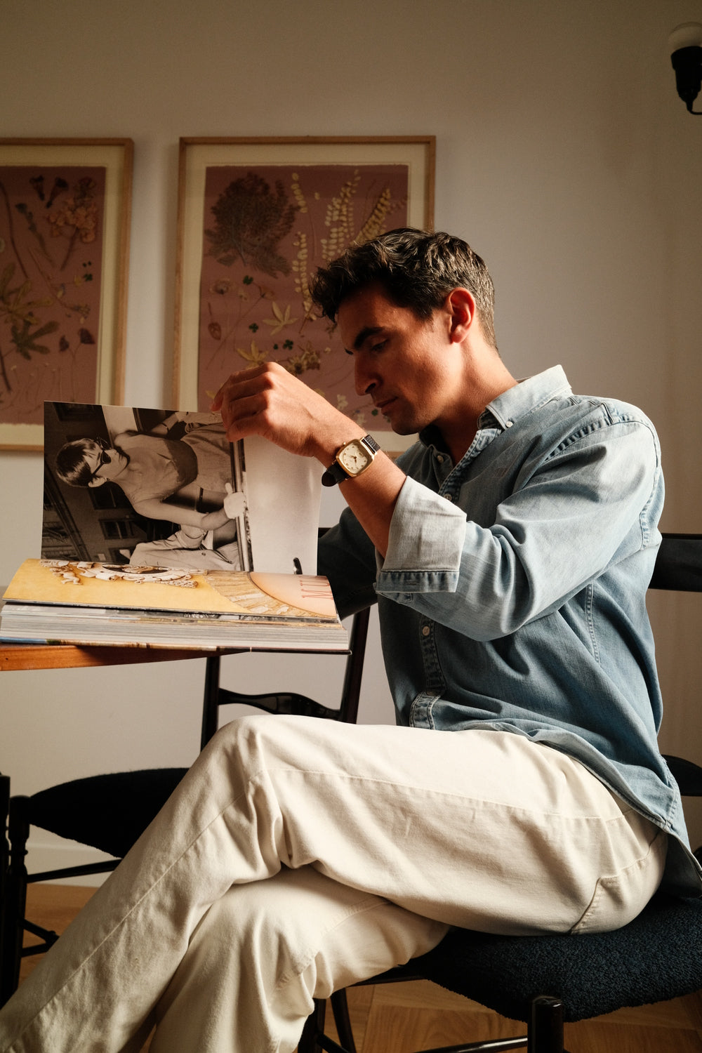 Man wearing denim shirt and AV86 watch with leather strap, sitting while browsing a large art book in softly lit room.