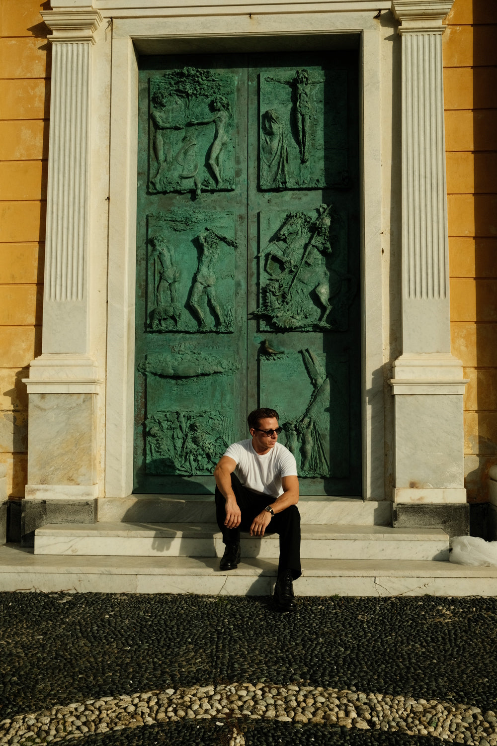 Man wearing black trousers and white t-shirt sitting on steps in front of large ornate bronze relief door with classical motifs