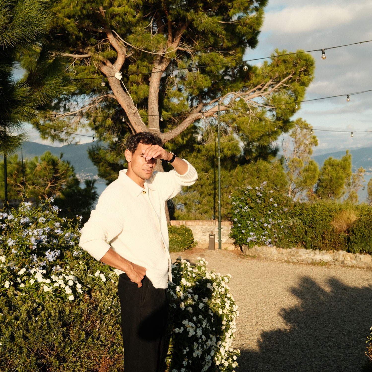 Man shielding eyes from sun standing in garden with pine trees, flowering shrubs, string lights, and distant mountain view.