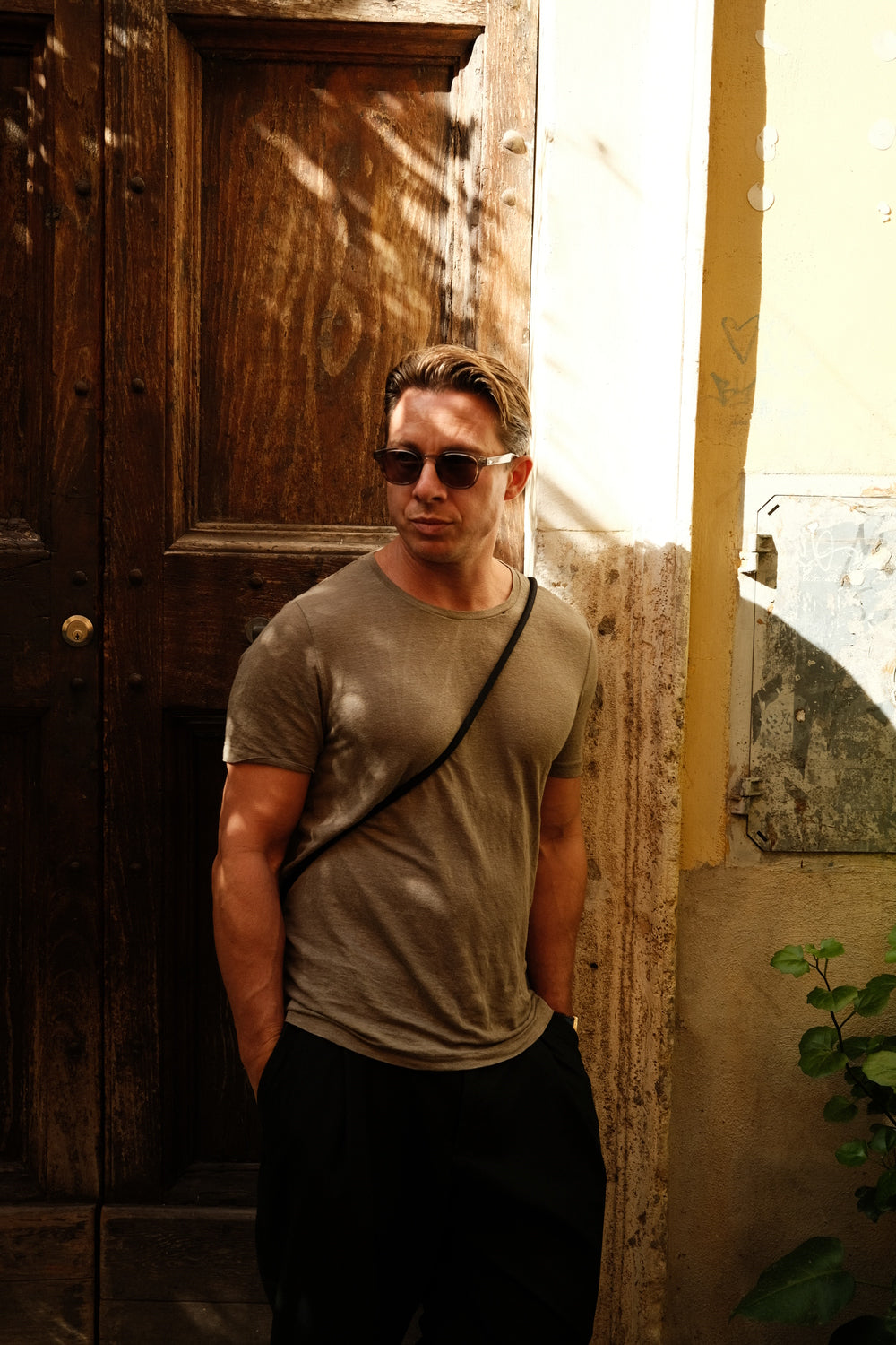 Man in casual minimalist outfit wearing 1954 Amalfi Mechanical, Gold / Midnight Blue by AV86 standing by rustic wooden door in warm sunlight