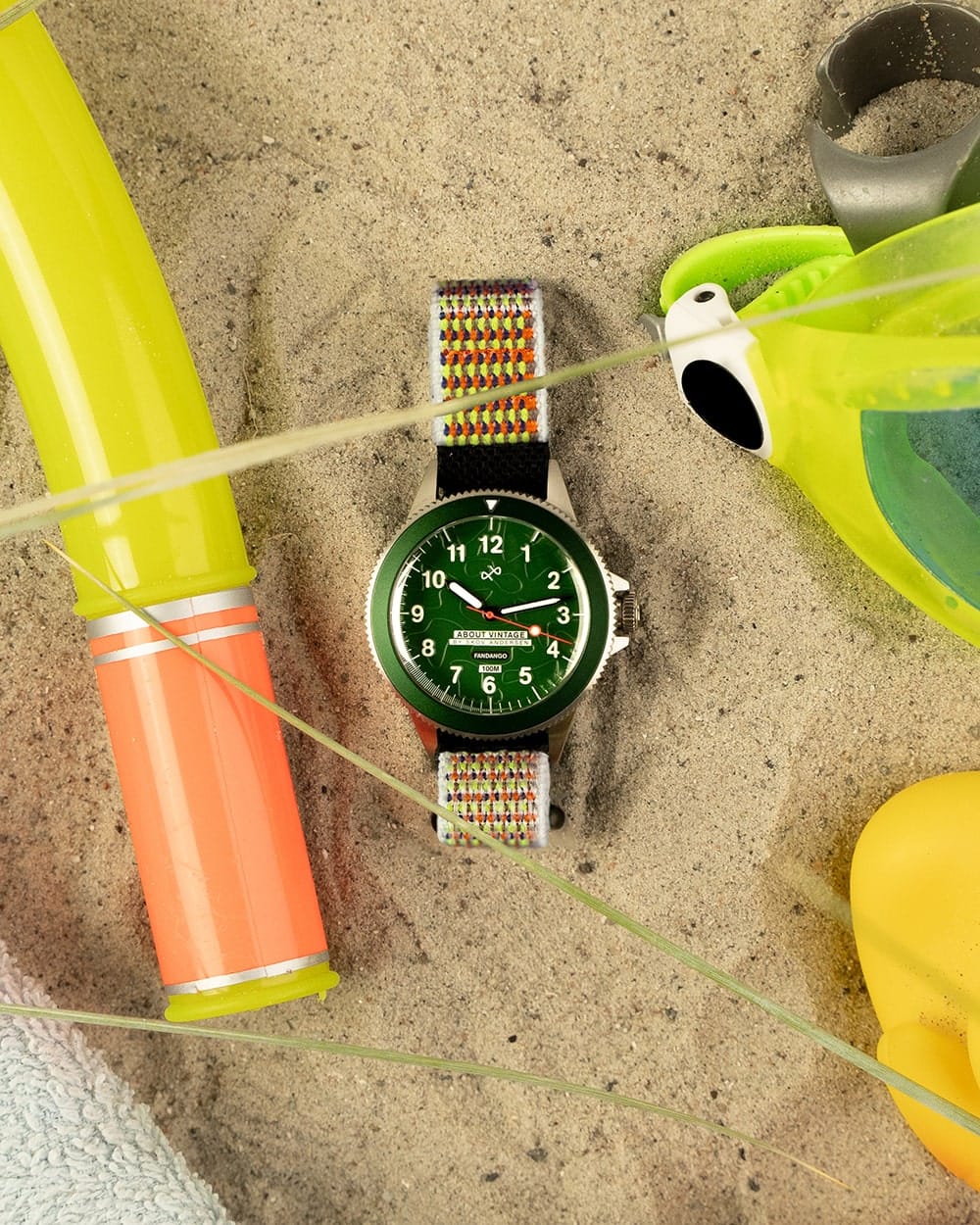 AV86 diver wristwatch with a green Arabic numeral dial and colorful woven strap resting on sand beside pool toys and goggles