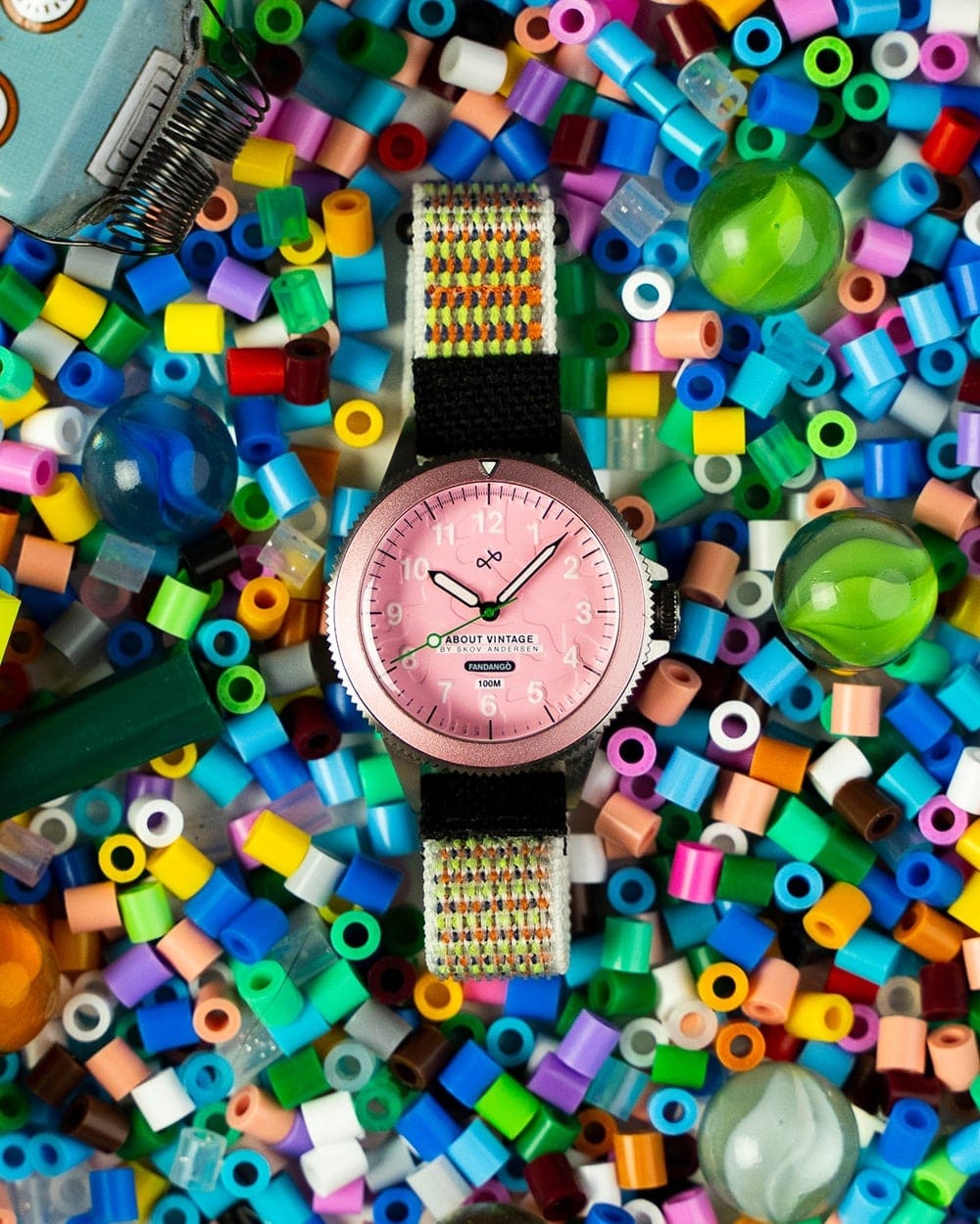 AV86 round wristwatch with pink Arabic numeral dial, black and multicolor woven strap, and polished case amid colorful beads