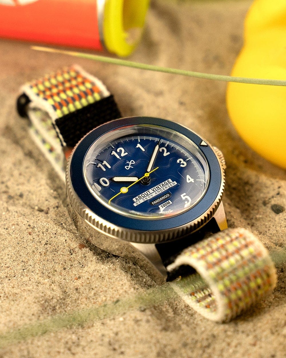 AV86 diver watch with blue Arabic numeral dial, rotating bezel and woven strap resting in sand beside colorful pool floats