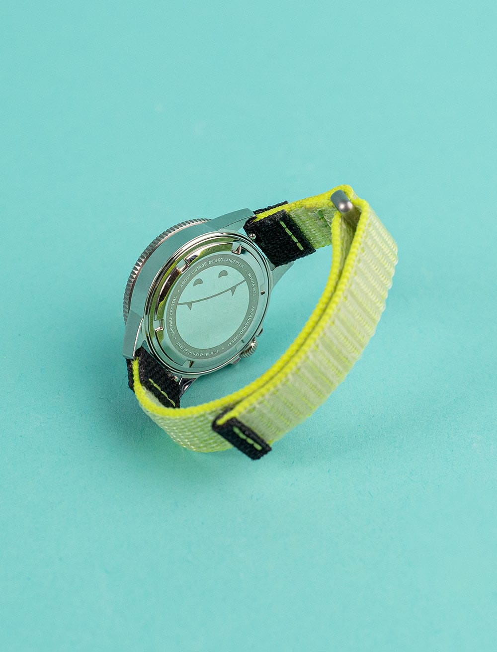 AV86 wristwatch shown from the polished caseback, featuring a round metal case and bright yellow woven strap on a turquoise background