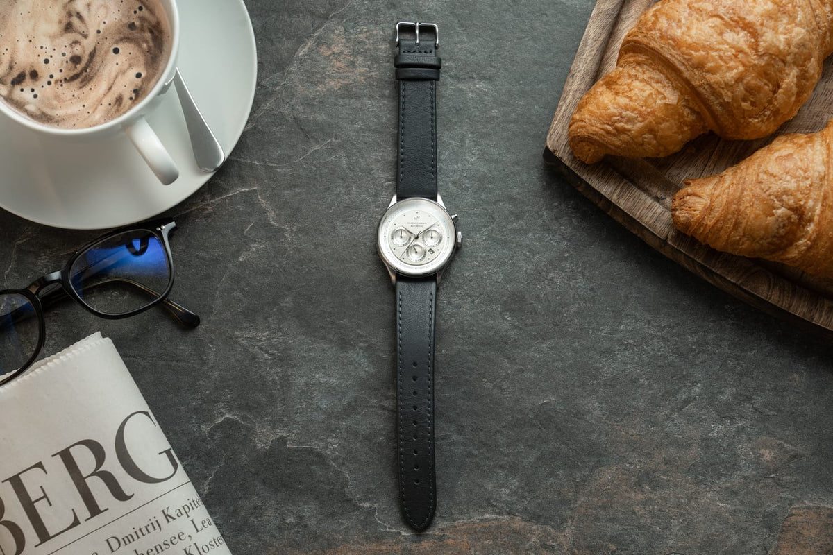 1564 Chronodate Automatic by AV86 features a vintage-inspired automatic chronograph with a refined white dial, multiple subdials, sapphire crystal, and a smooth black leather strap, exemplifying Danish horological craftsmanship and Nordic minimalism, styled alongside a breakfast setting with coffee, croissants, and a newspaper.