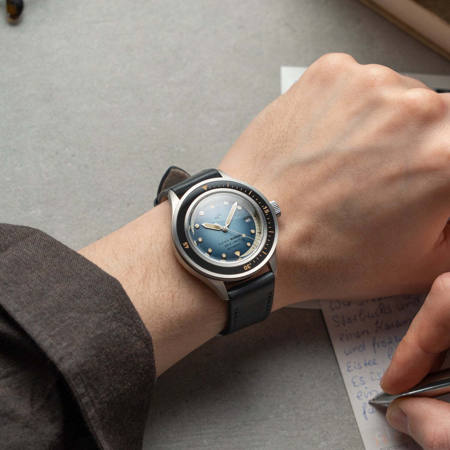 1956 Marine Automatic, Steel / Blue Fade wristwatch by AV86 with a blue fade dial and black leather strap on a wrist writing notes