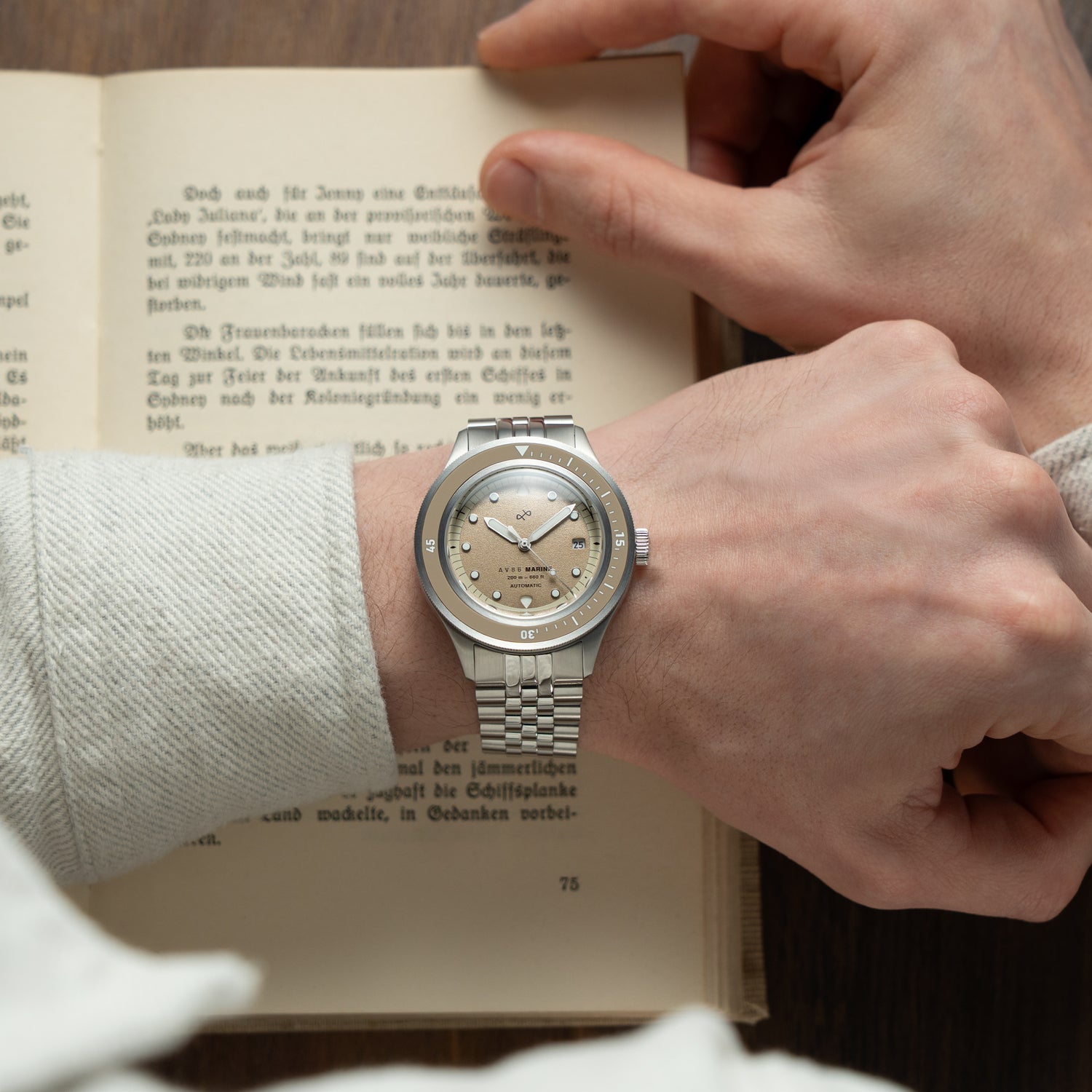 1956 Marine Automatic, Steel / Sand wristwatch by AV86 with vintage-inspired dial and stainless steel bracelet on a wrist over an open book